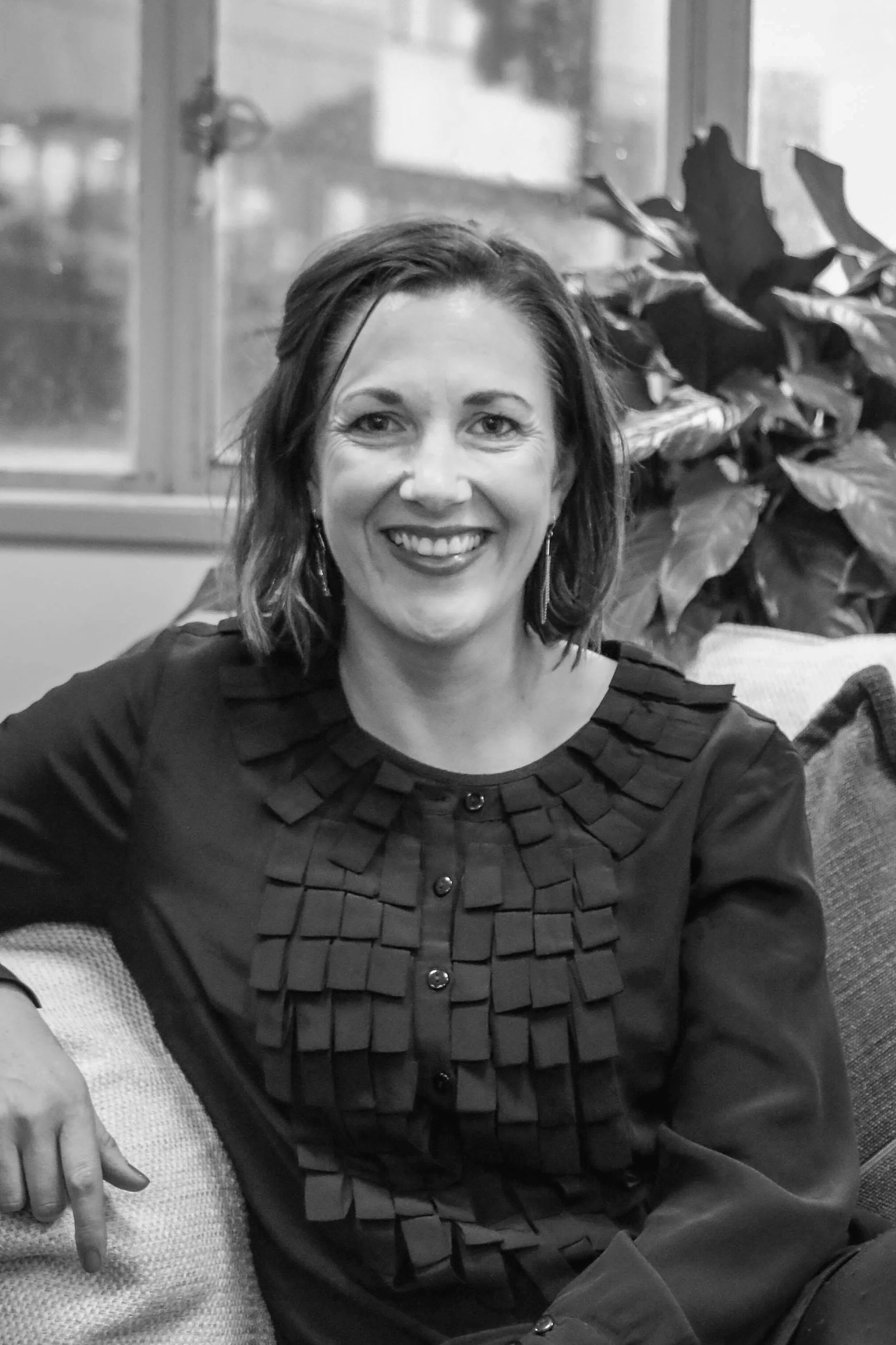 Black and white photo of a woman with shoulder-length hair smiling at the camera, wearing a dark top with decorative pleats, sitting on a couch with large leafy plants and windows in the background.