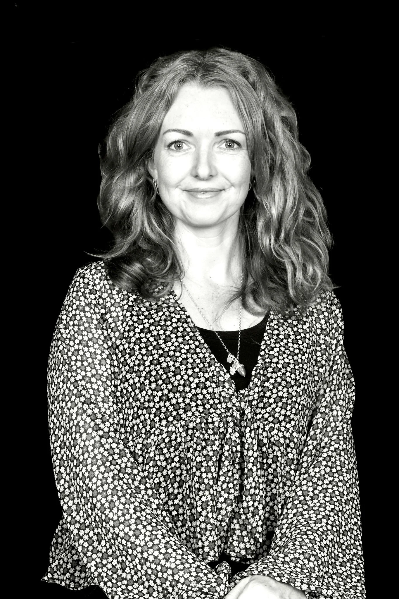 Black and white portrait of a woman with wavy shoulder-length hair, wearing a floral patterned blouse and smiling.