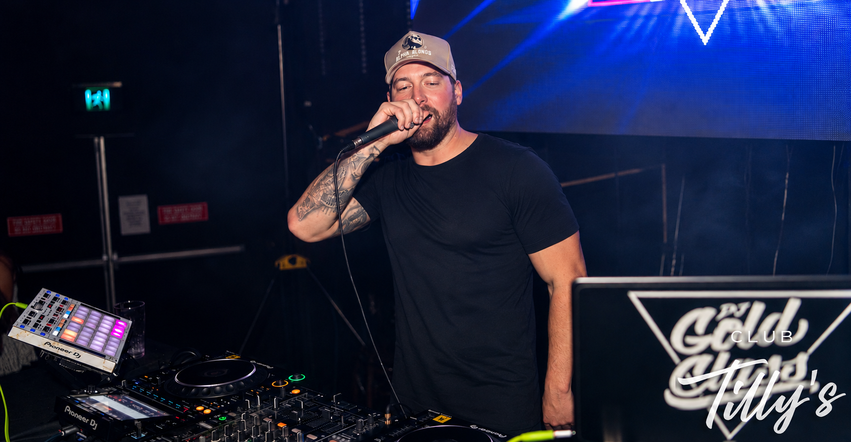 A man with tattoos on his arm holding a microphone and DJ equipment in a nightclub. He is wearing a black t-shirt and a beige cap. The background features a large digital screen with blue lighting and a logo that reads 'The Club Tilly's'.