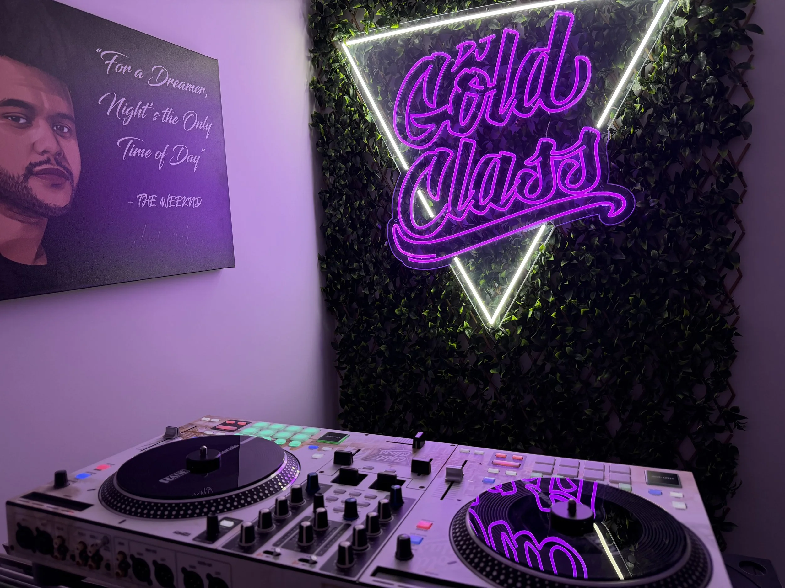 A DJ setup with turntables and a mixer on a table, a neon sign that says 'good vibes' on a green leafy wall, and a poster with a quote and a portrait of a young man.