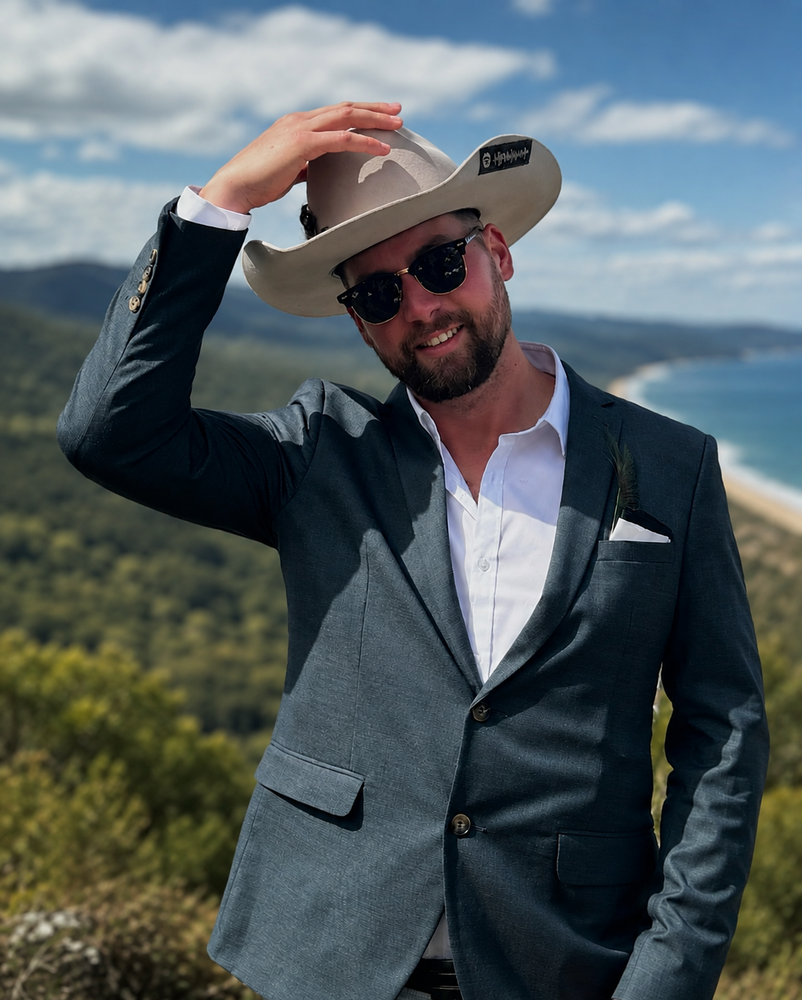 A man in a suit wearing sunglasses and a cowboy hat, smiling outdoors with a coastal landscape and blue sky in the background.