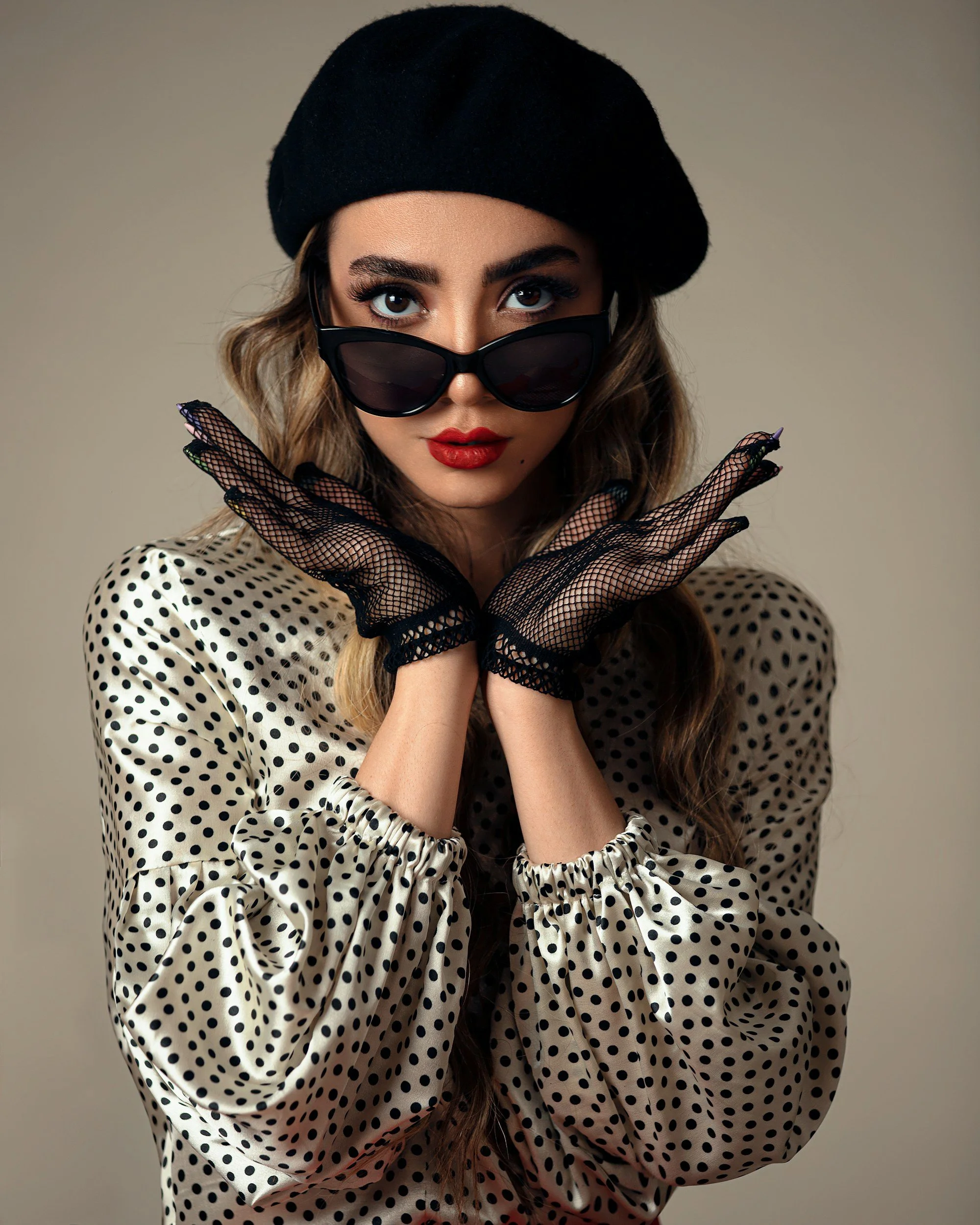 A woman with long, wavy hair wearing black sunglasses, a black beret, a polka dot blouse, and black fishnet gloves, posing with crossed arms and a confident expression.