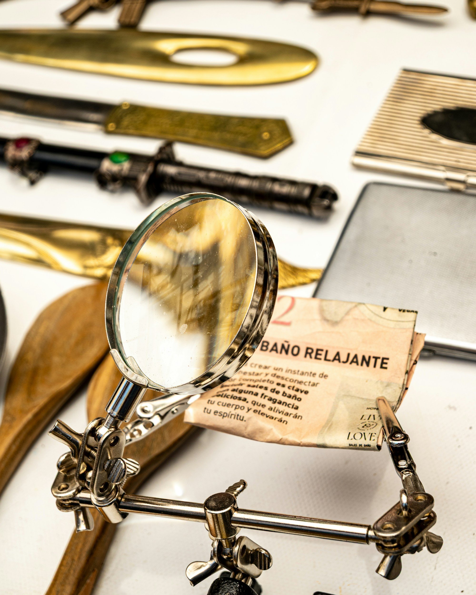 Close-up of a small metal figurine of a person, using a magnifying glass to examine a pamphlet titled 'Baño Relajante' on a white surface, surrounded by tools and objects.