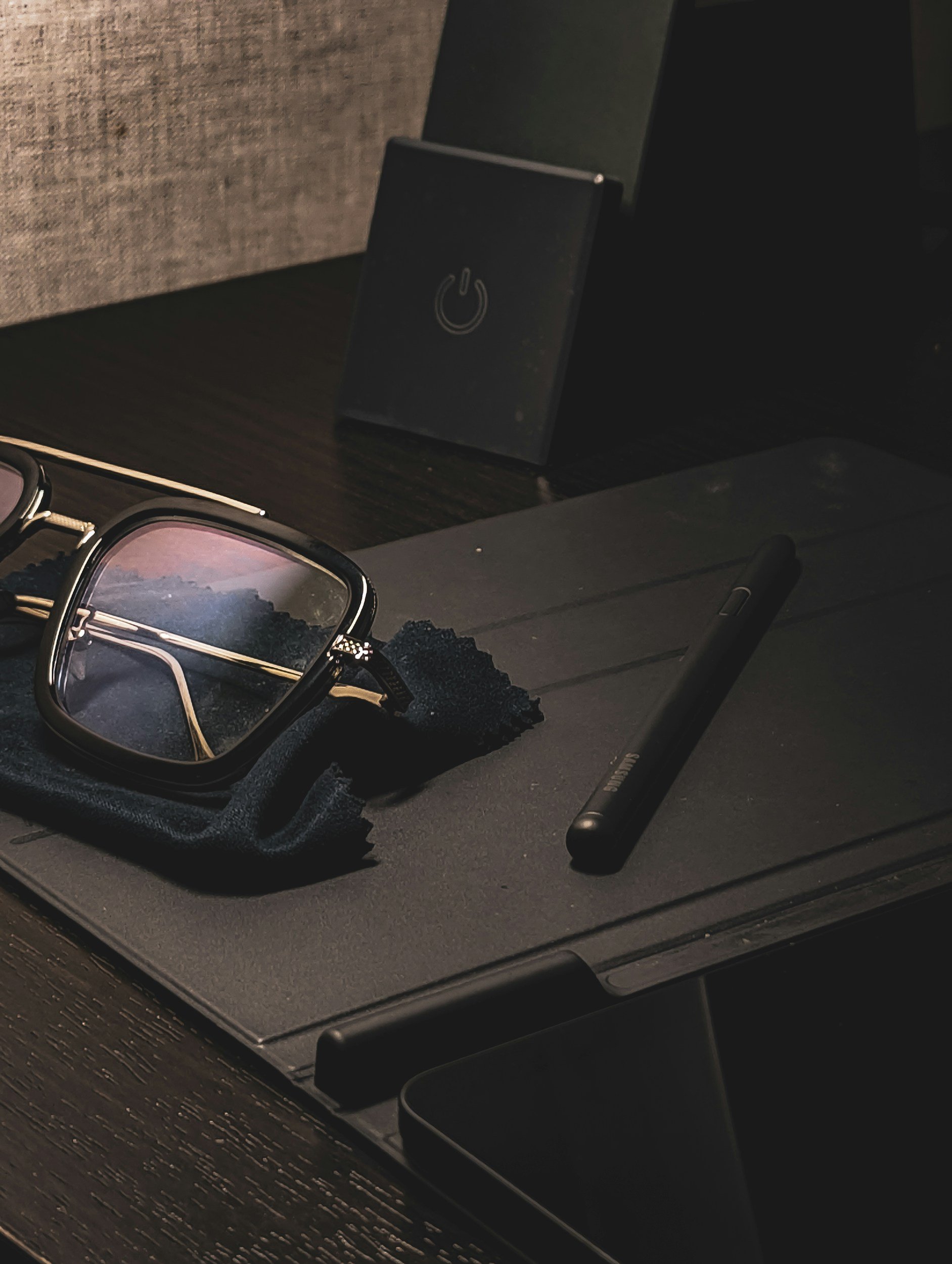 A pair of eyeglasses with a metal frame, a black device with a power button icon, a black pen, and a black case on a dark textured surface.