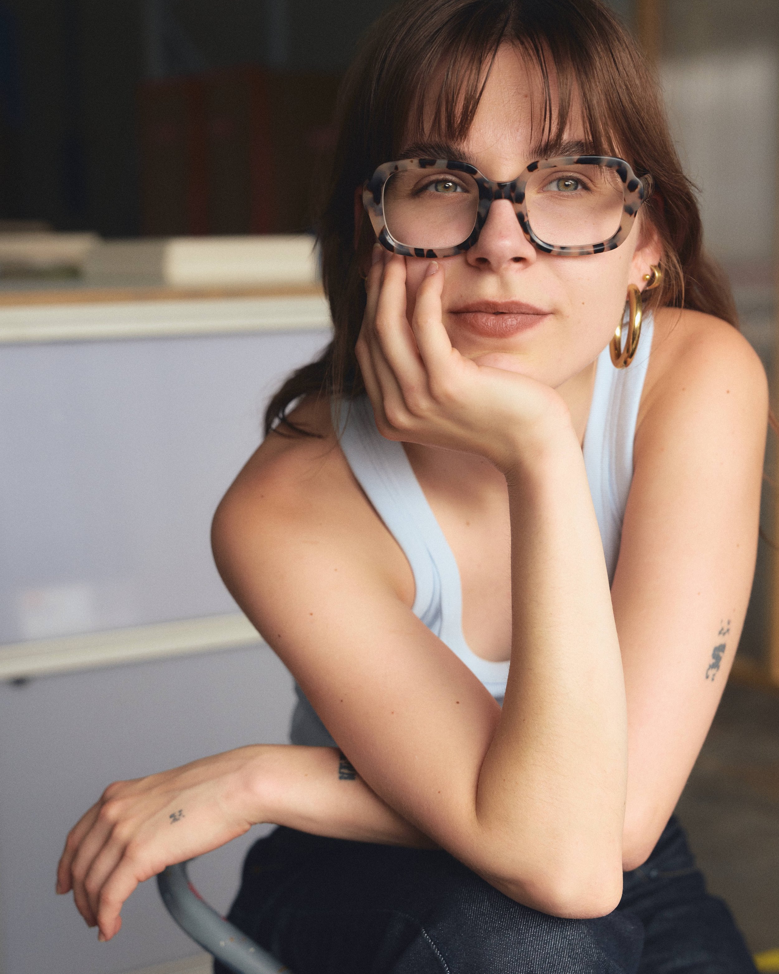 A woman sitting with her hand on her chin, wearing large tortoiseshell glasses, gold hoop earrings, and a white sleeveless top.