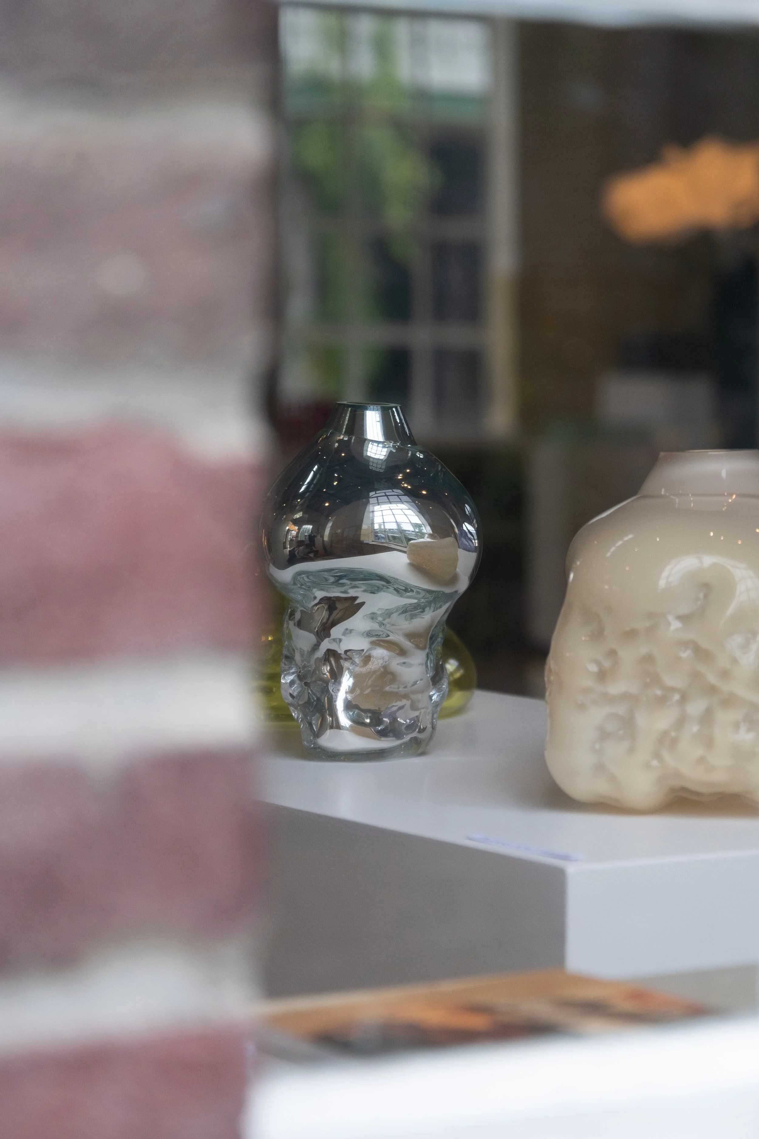 A reflective silver-colored decorative object resembling a skull and a white object with a textured surface is on display on a white surface. In the background, there is a blurred interior space with a window, shelves, and green plants.
