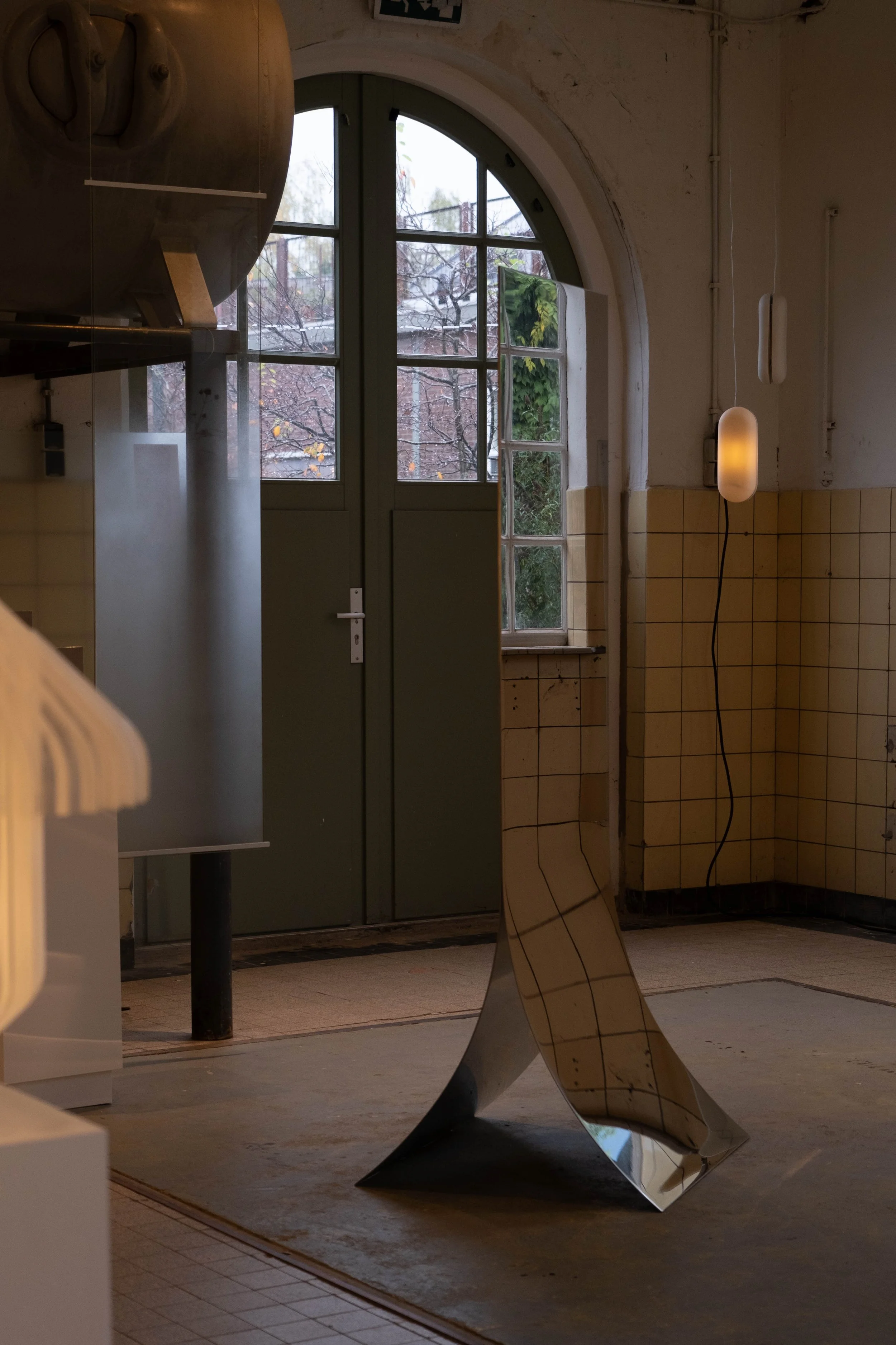 Inside a room with large arched window, a metal sculpture of wavy form, a suspended cylindrical lamp, and a tiled wall. Outside the window are bare trees and a cloudy sky.