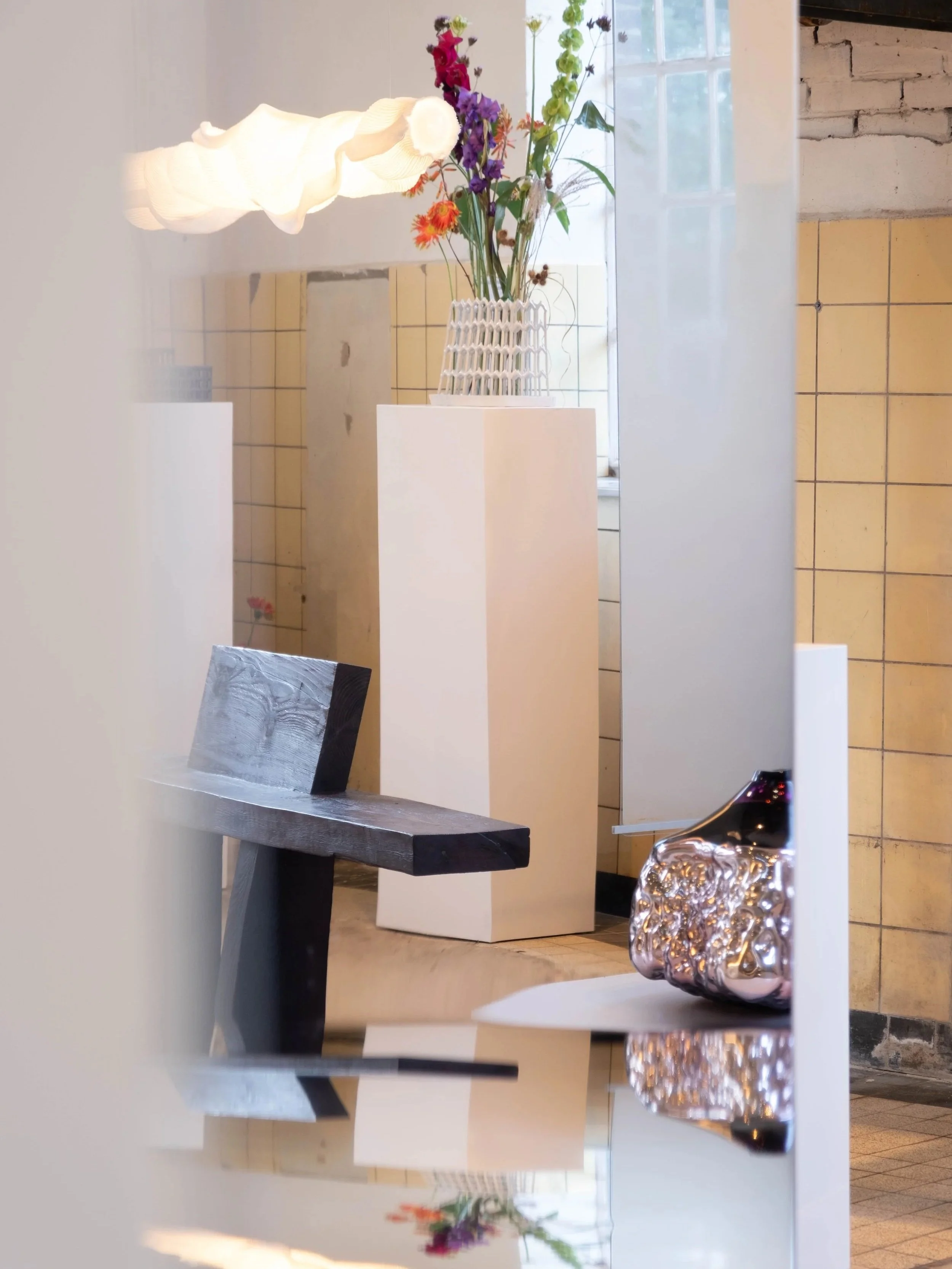 A space with a floral arrangement in a white vase on a white pedestal, a small side table, and a modern white hanging lamp. The background includes a wall with yellow tiles, a window with daylight, and a portion of a brick wall.