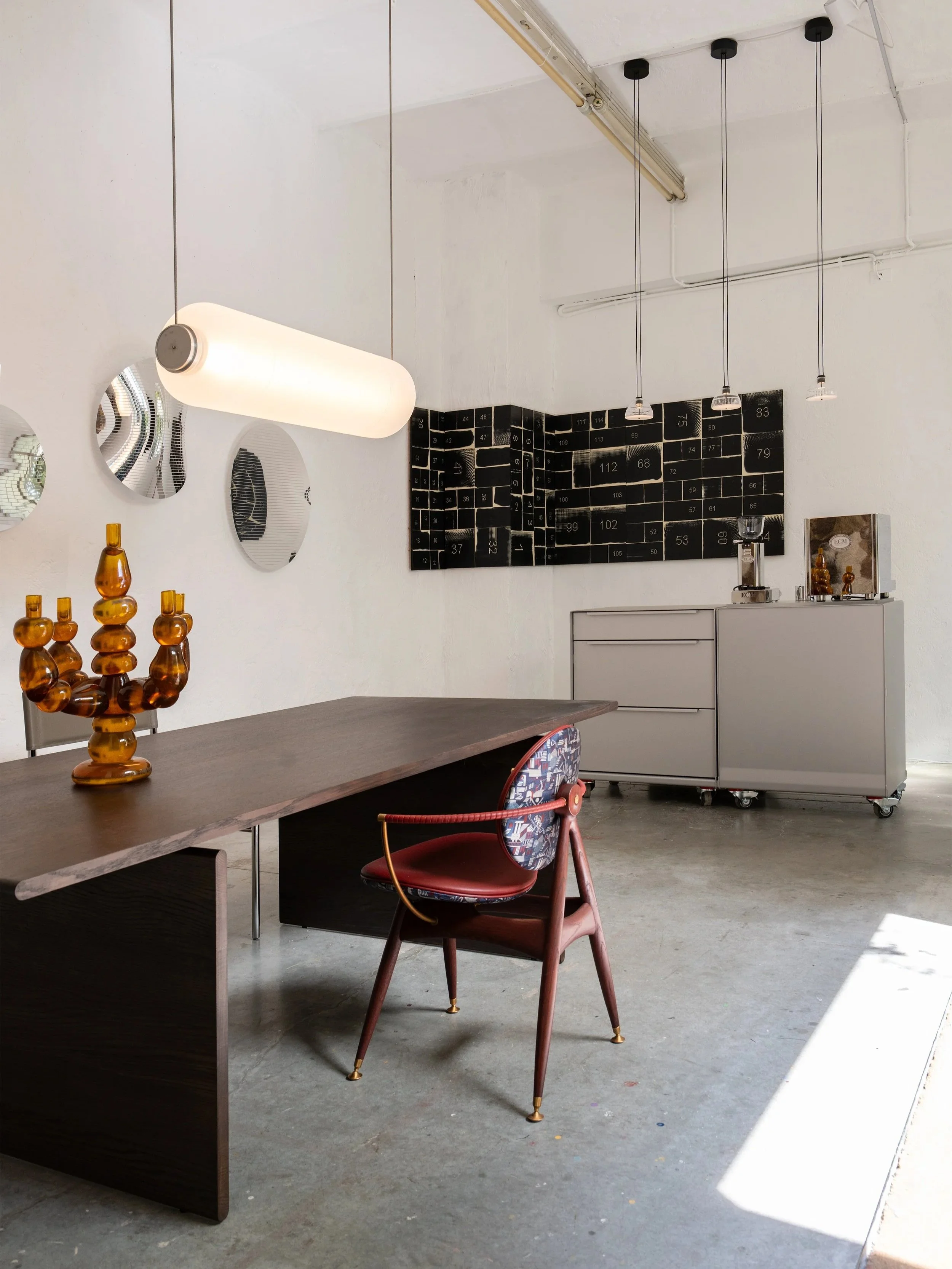 Interior view of a modern, minimalistic cafe or workspace with a wooden table, a red chair with patterned upholstery, a decorative amber glass sculpture, a black dashboard-style wall art, hanging ceiling lights, and a white cabinet with small decorative items on it.