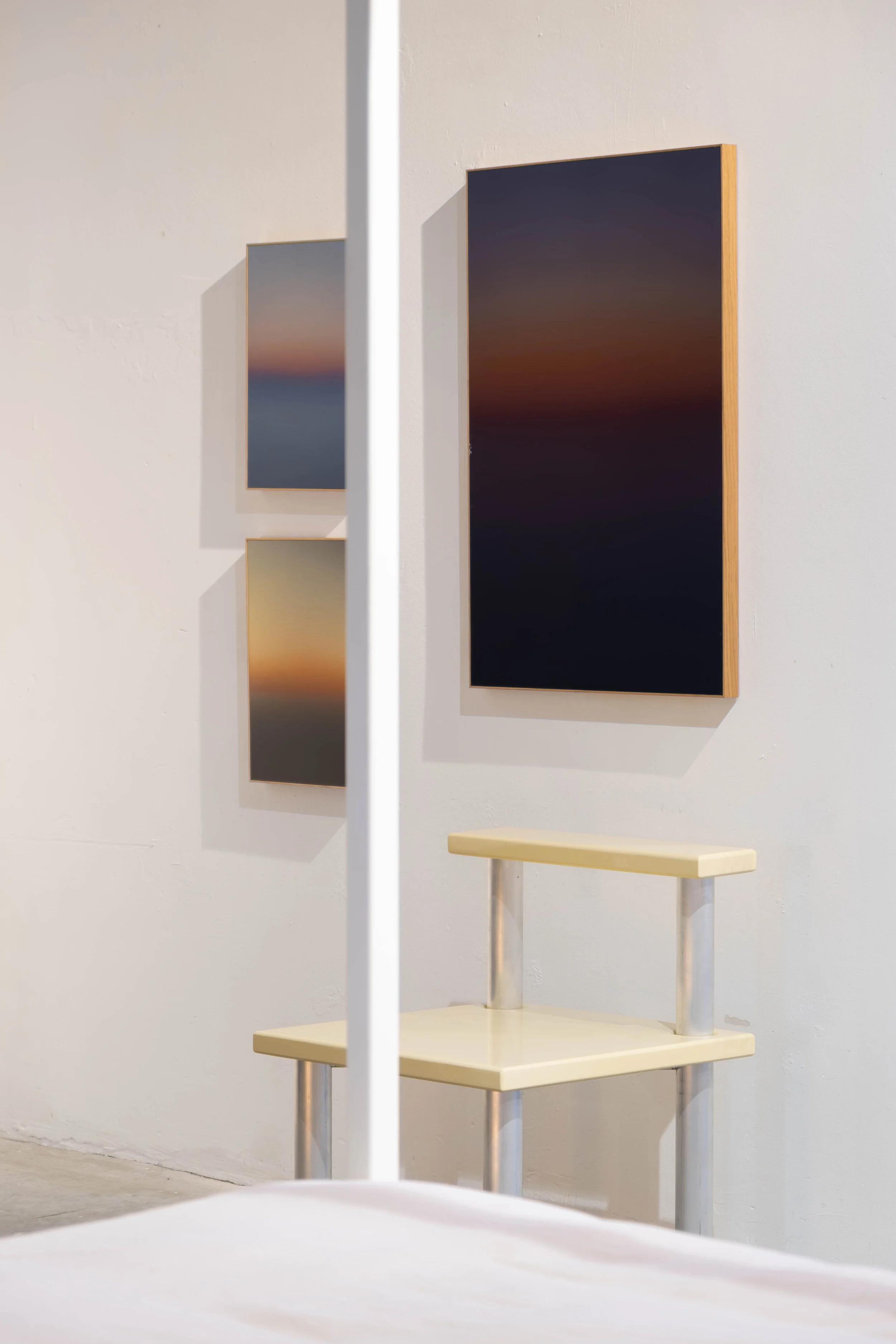 An art gallery with two framed landscape photographs on a white wall and a beige table with metal legs in front of them. The photographs depict sunset or sunrise scenery with dark silhouettes and colorful skies.