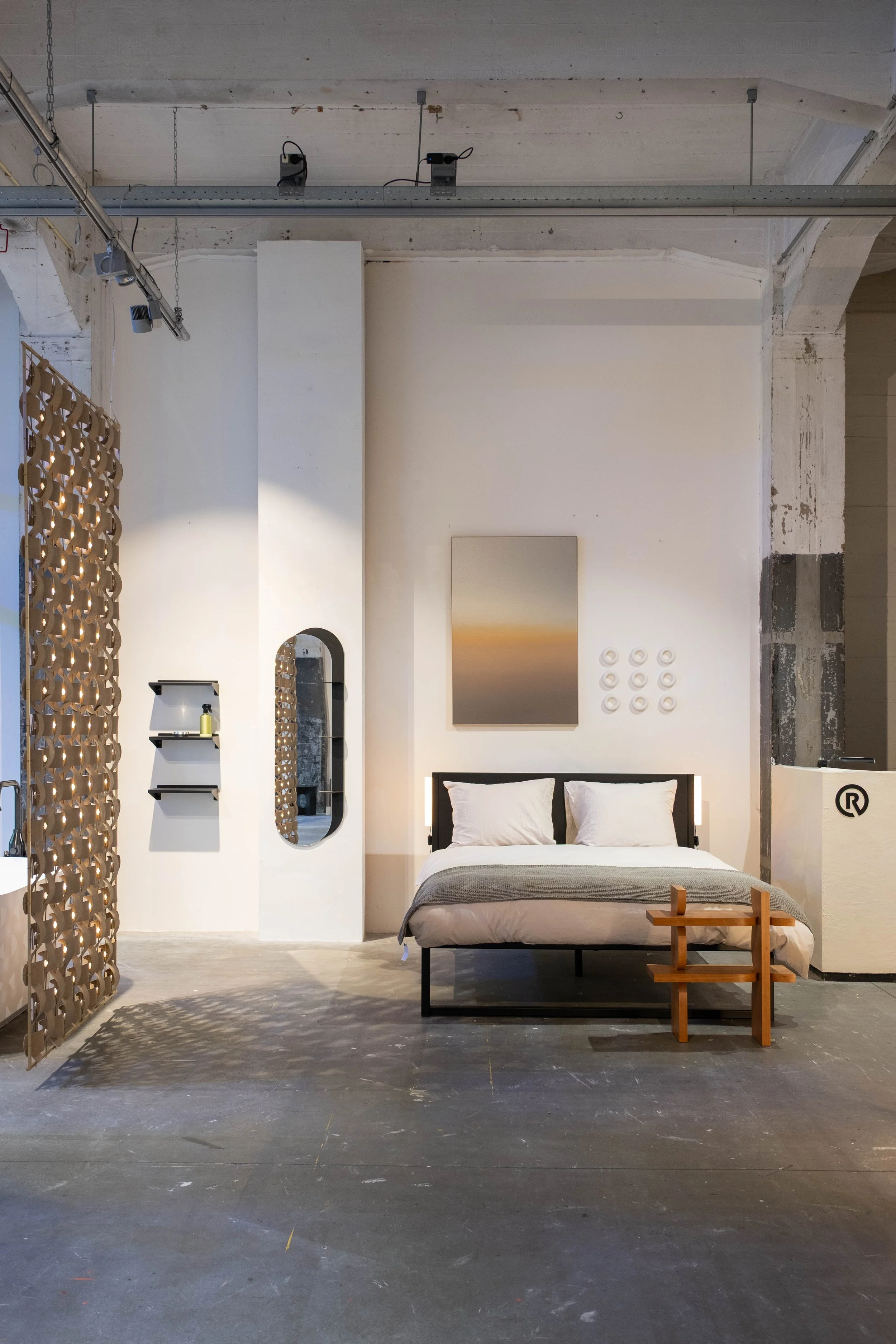 Interior of a modern, minimalist bedroom with a low bed, a small wooden stool, and a textured wall art piece, in an industrial-style space with exposed concrete and ceiling fixtures.