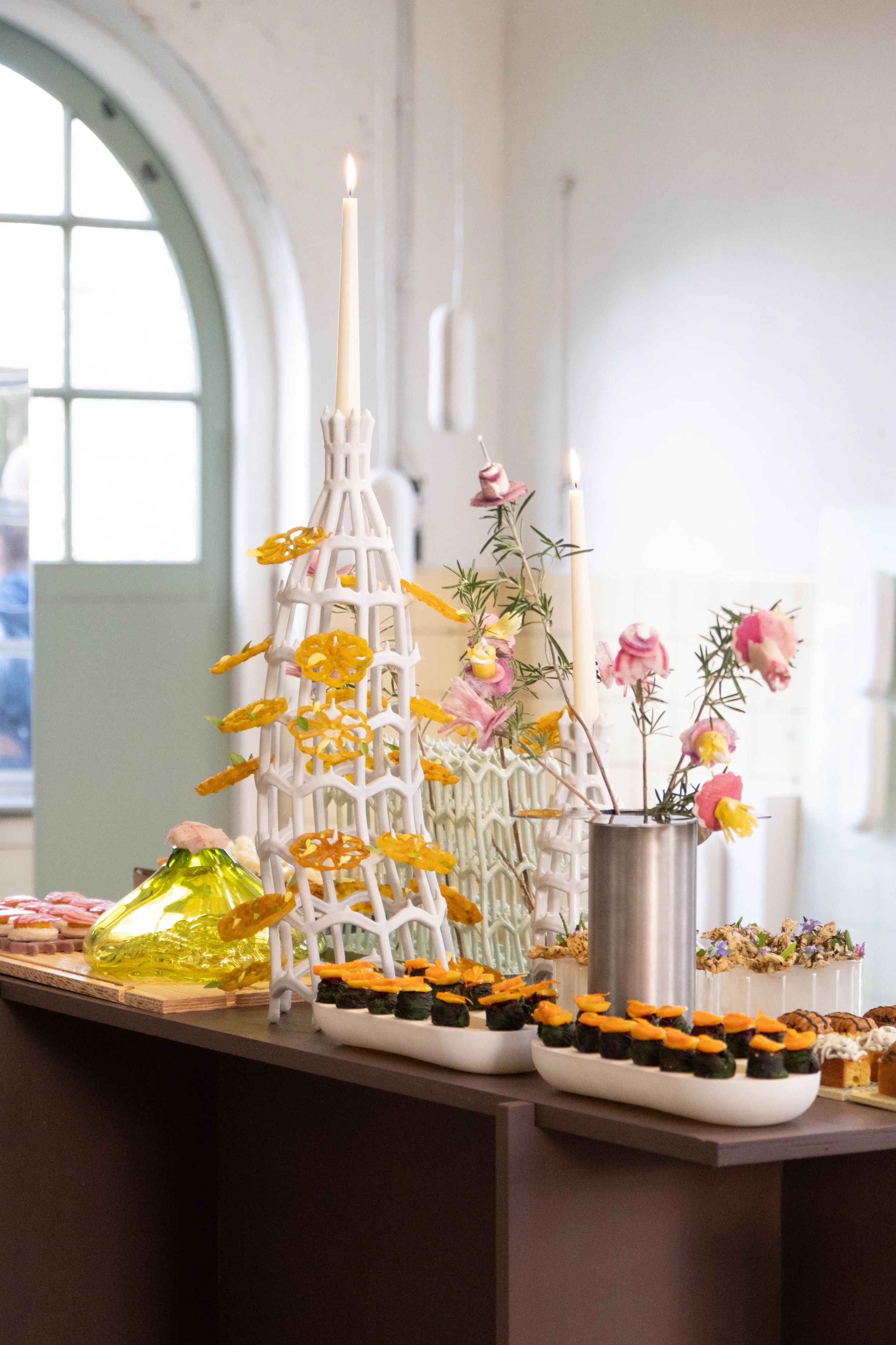 Display of various beautiful and assorted desserts, including cupcakes, cookies, and candies, with a decorative white structure, candles, and floral arrangements on a table.