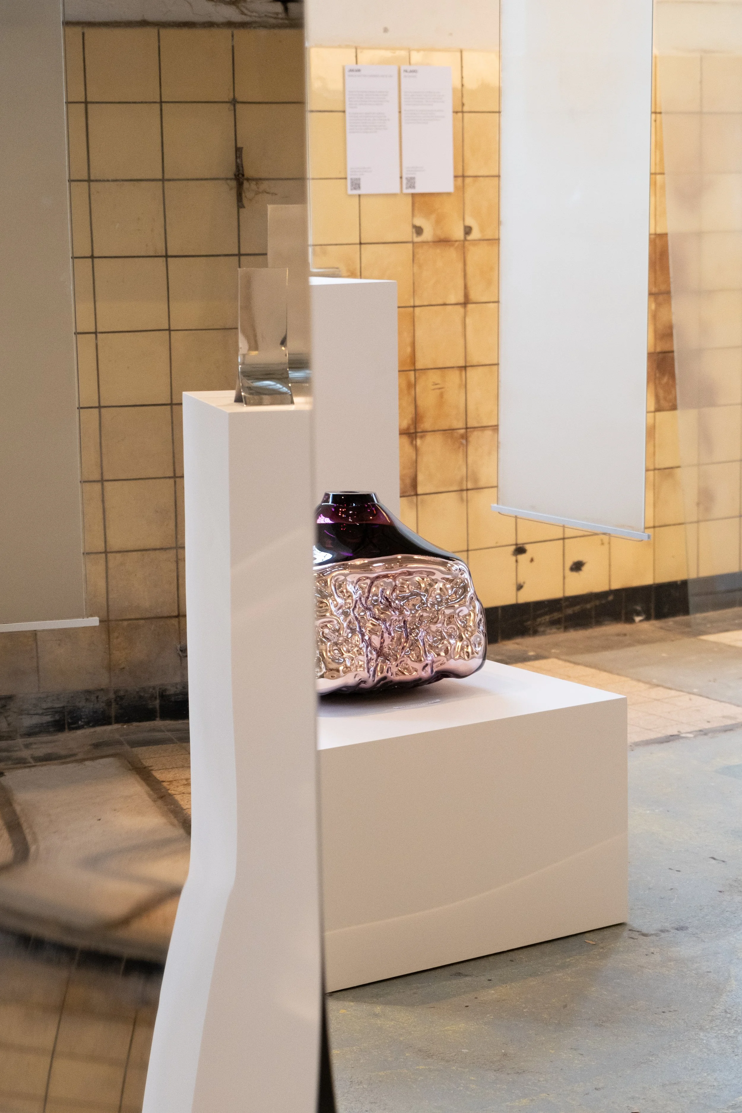 A gallery wall with a white pedestal displaying a shiny, textured, metallic sculpture and a smaller, smooth, dark-colored vase. In the background, beige and brown tiled walls with displayed papers or posters.