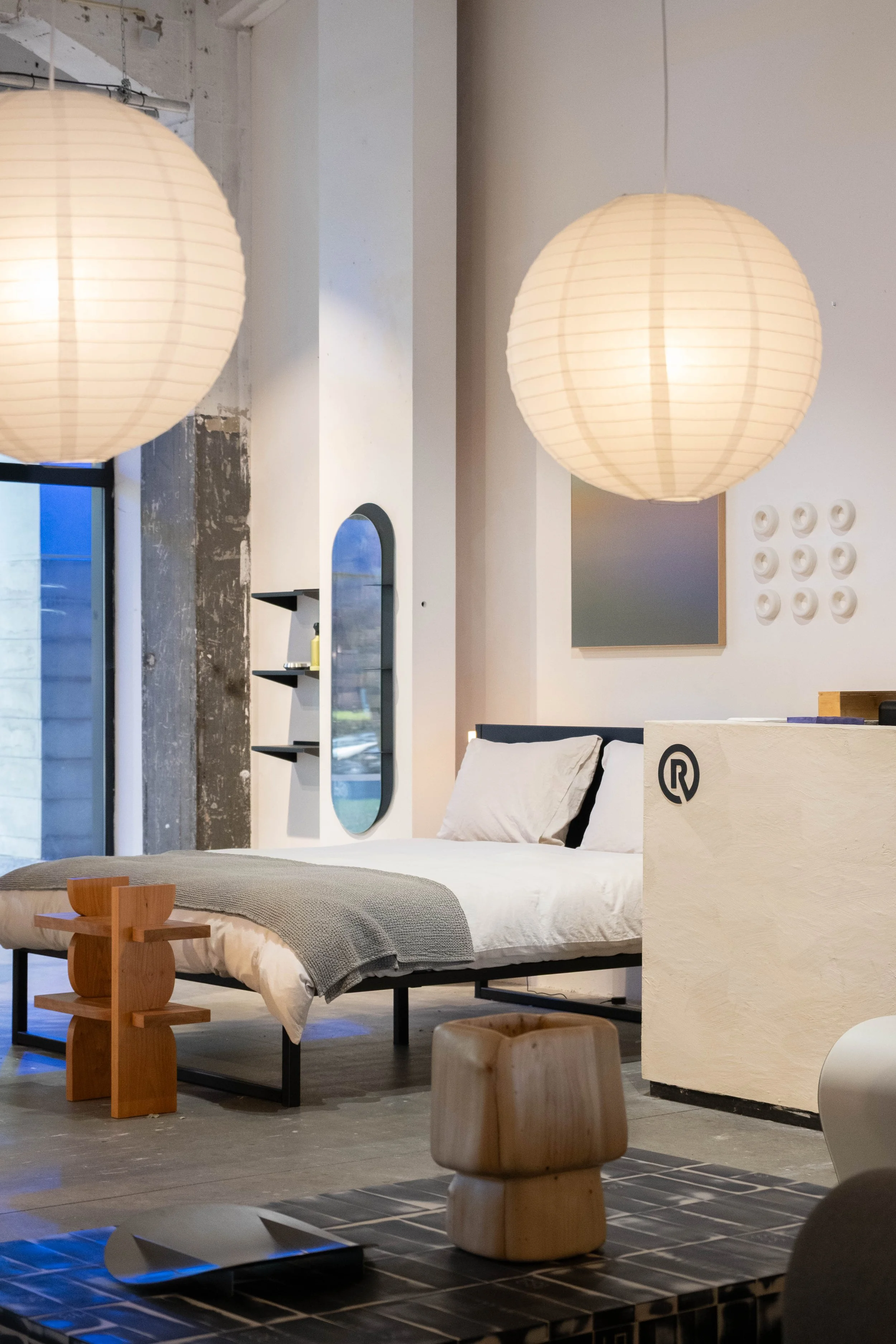 Interior of a modern furniture store with a bed, a small wooden side table, large hanging paper lantern lights, and minimalist decor elements.