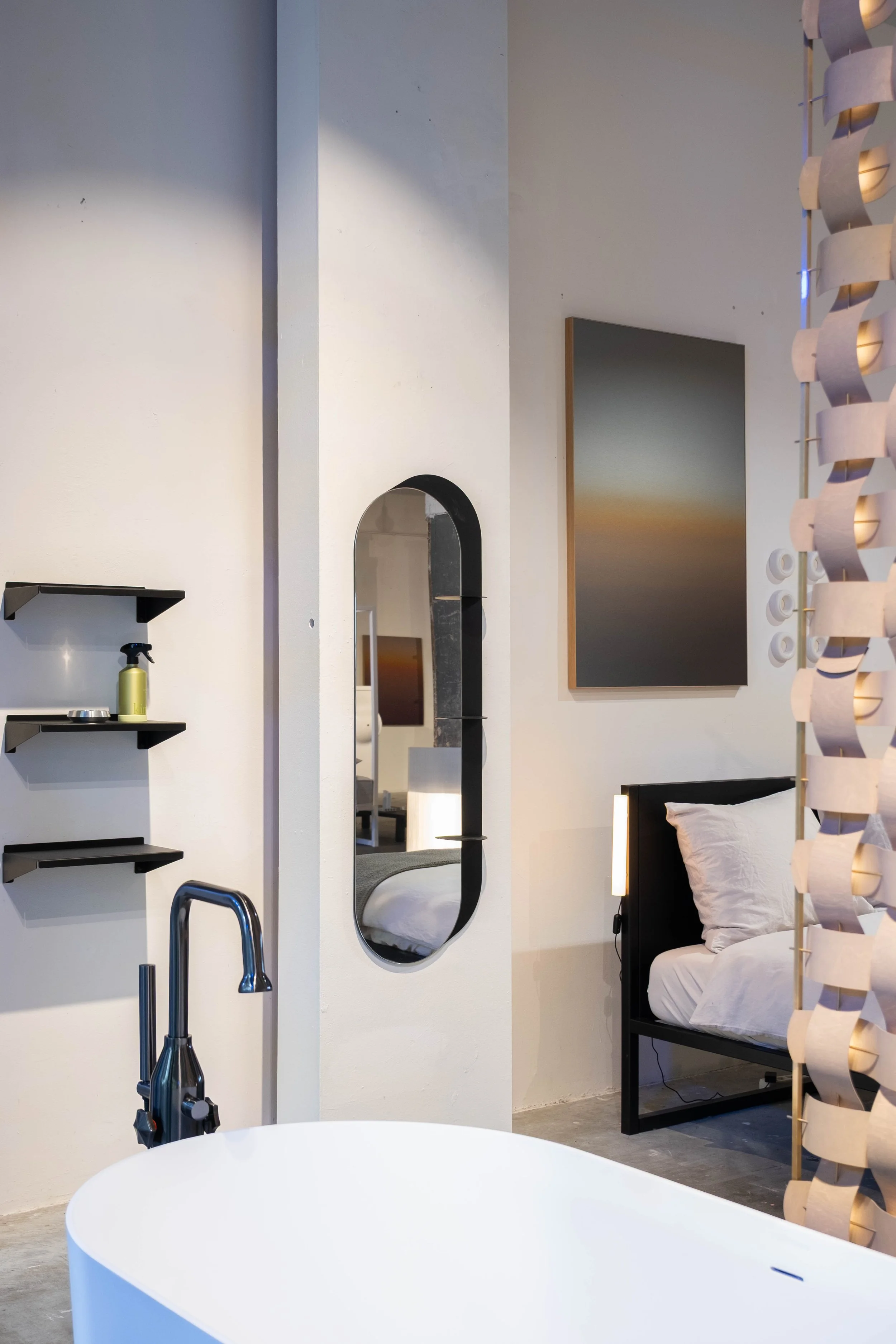 Modern interior space with a white bathtub in the foreground, a wall-mounted black shelf with a spray bottle and containers, an elongated oval mirror on a white wall, a bed with white bedding, and decorative elements including a standing sculpture made of connected wooden slats and a rectangular wall art piece depicting a sunset.