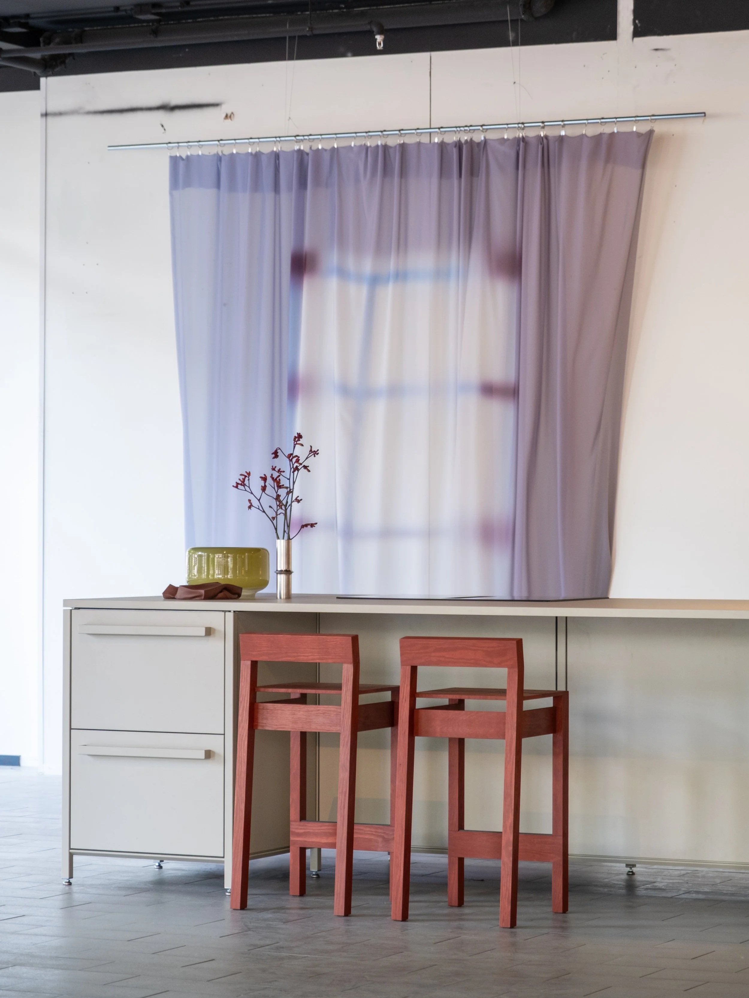 A dining area with a white cabinet, two red wooden chairs, a vase with a branch, a mustard-colored bowl, and purple curtains hanging behind the cabinet.