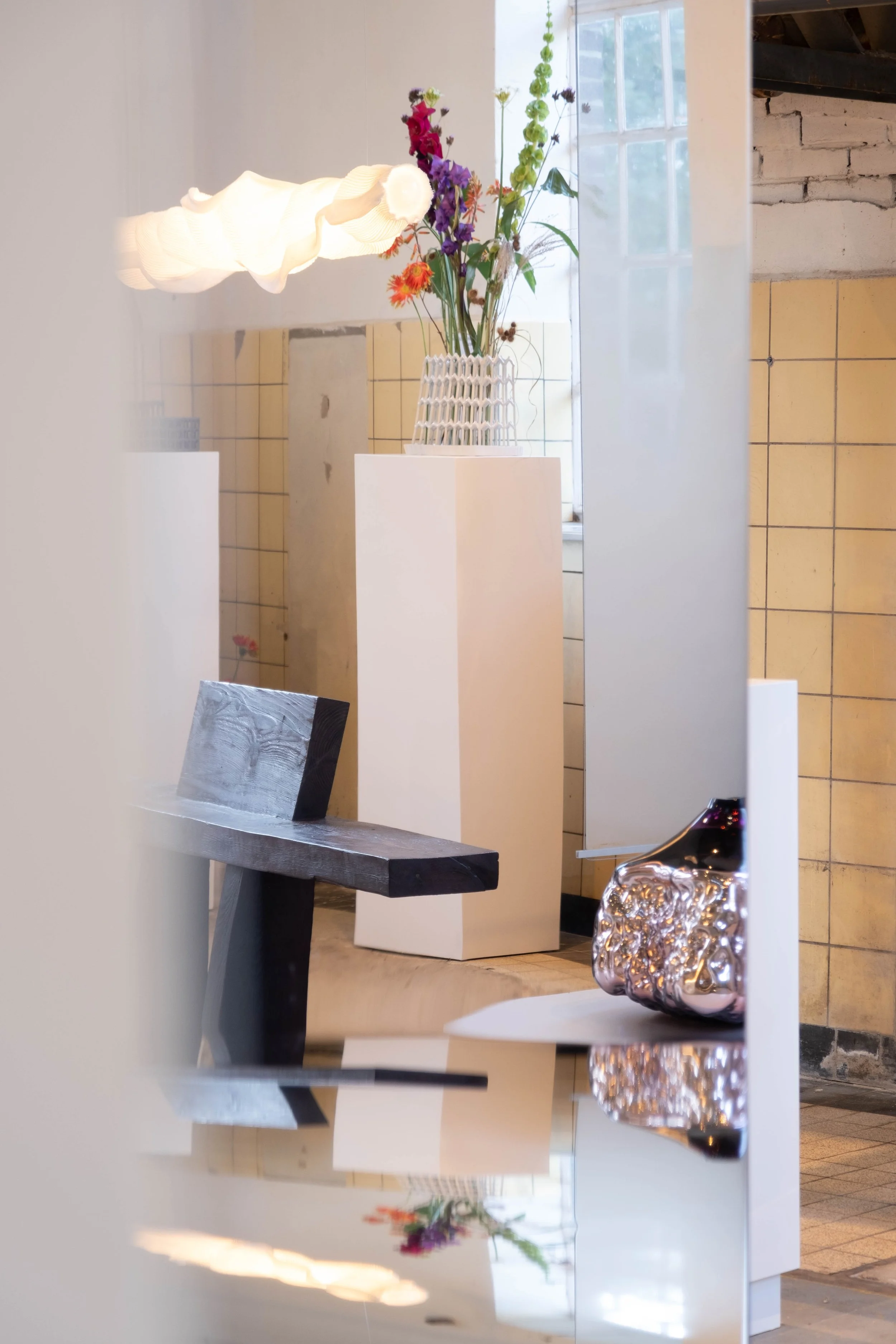 A jewelry store display with a glass vase holding a colorful bouquet of flowers, a white pedestal, and a black chair nearby. The background shows a window and a brick wall.