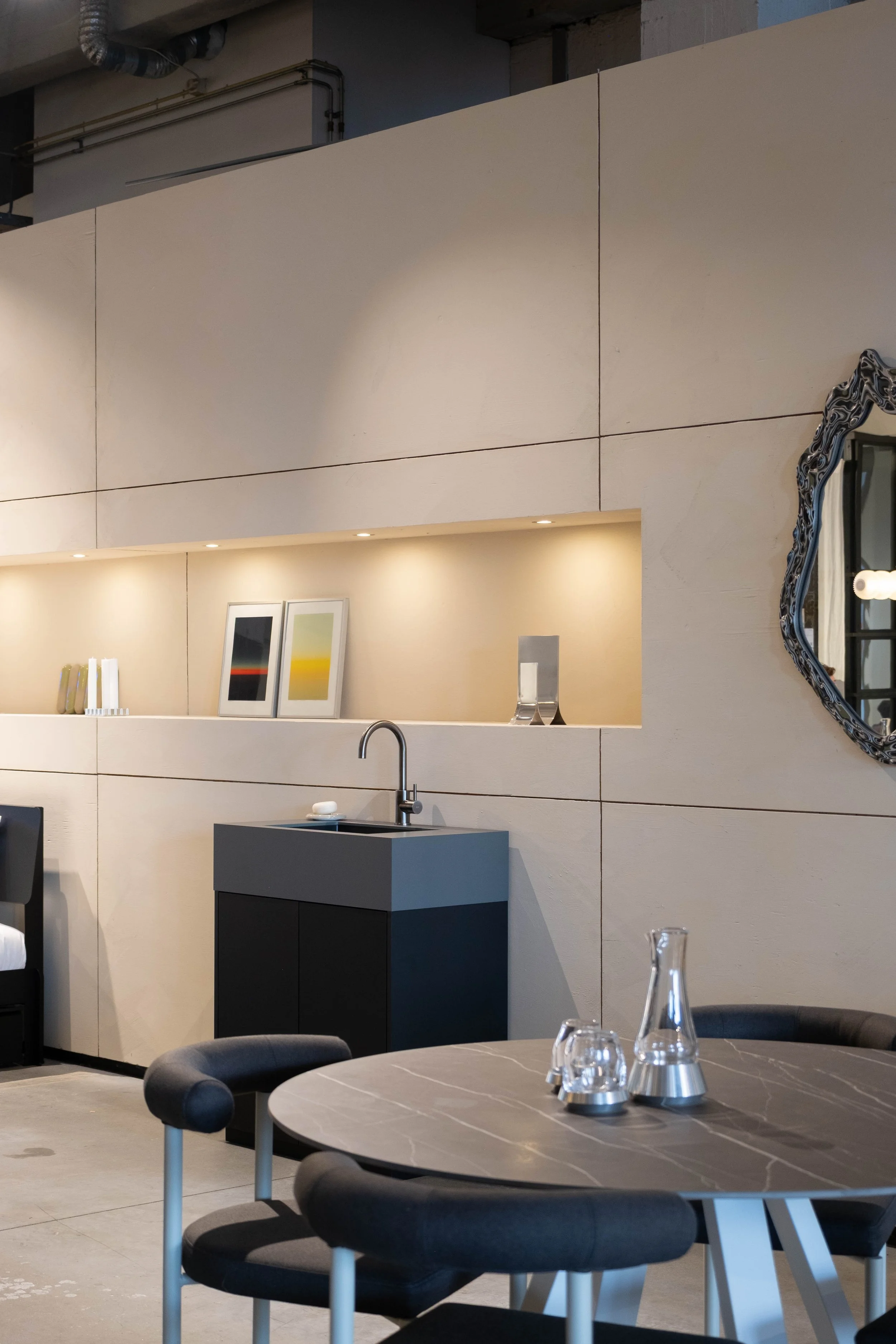 Modern dining area with round marble table, black chairs, decorative glassware, a black cabinet with a sink, and minimalist wall art, illuminated by built-in ceiling lights and natural light from a window.
