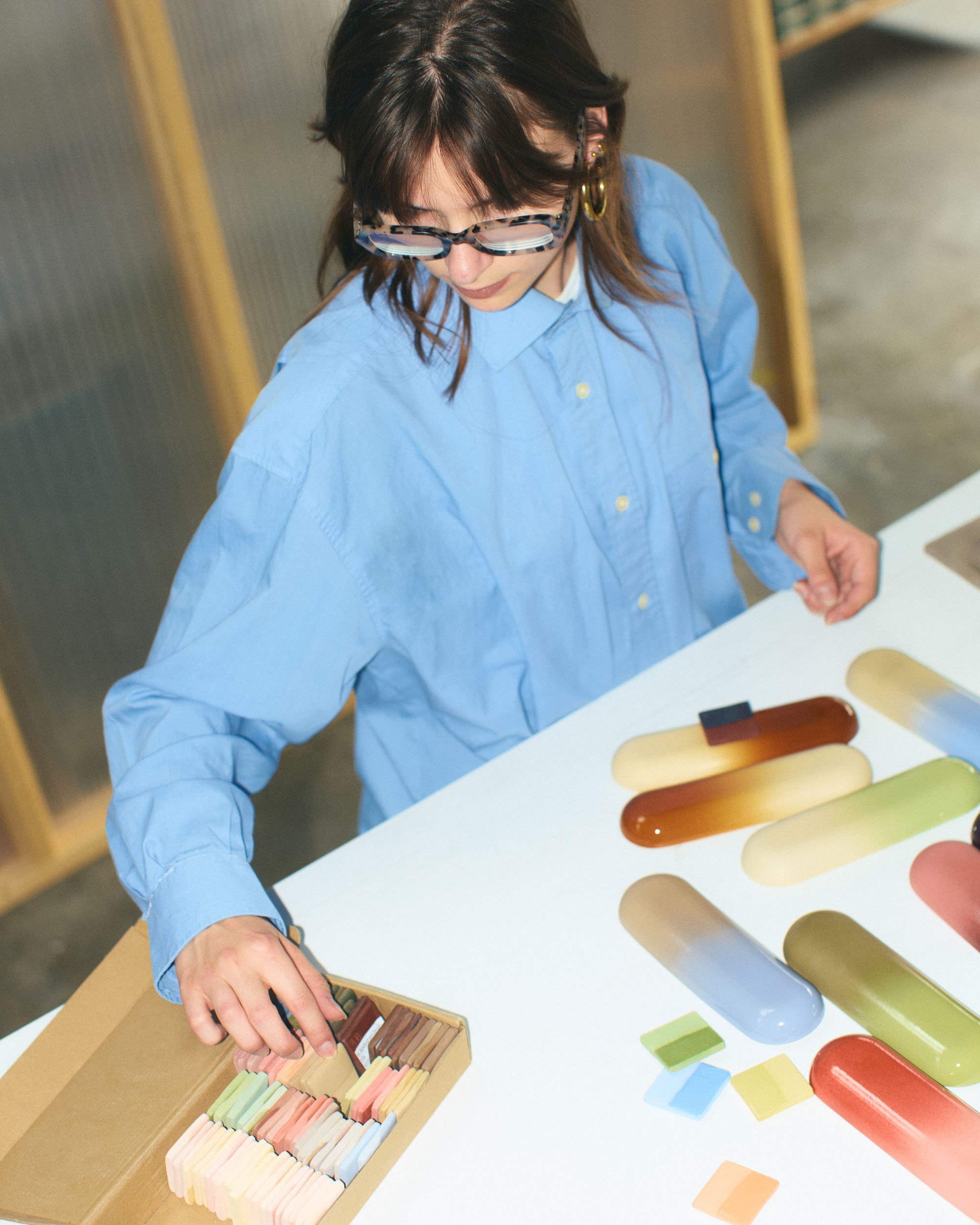 A woman wearing glasses and a blue shirt is at a table with ceramic pills, selecting a piece from a wooden box. On the table, there are large chalk sticks arranged in a row.