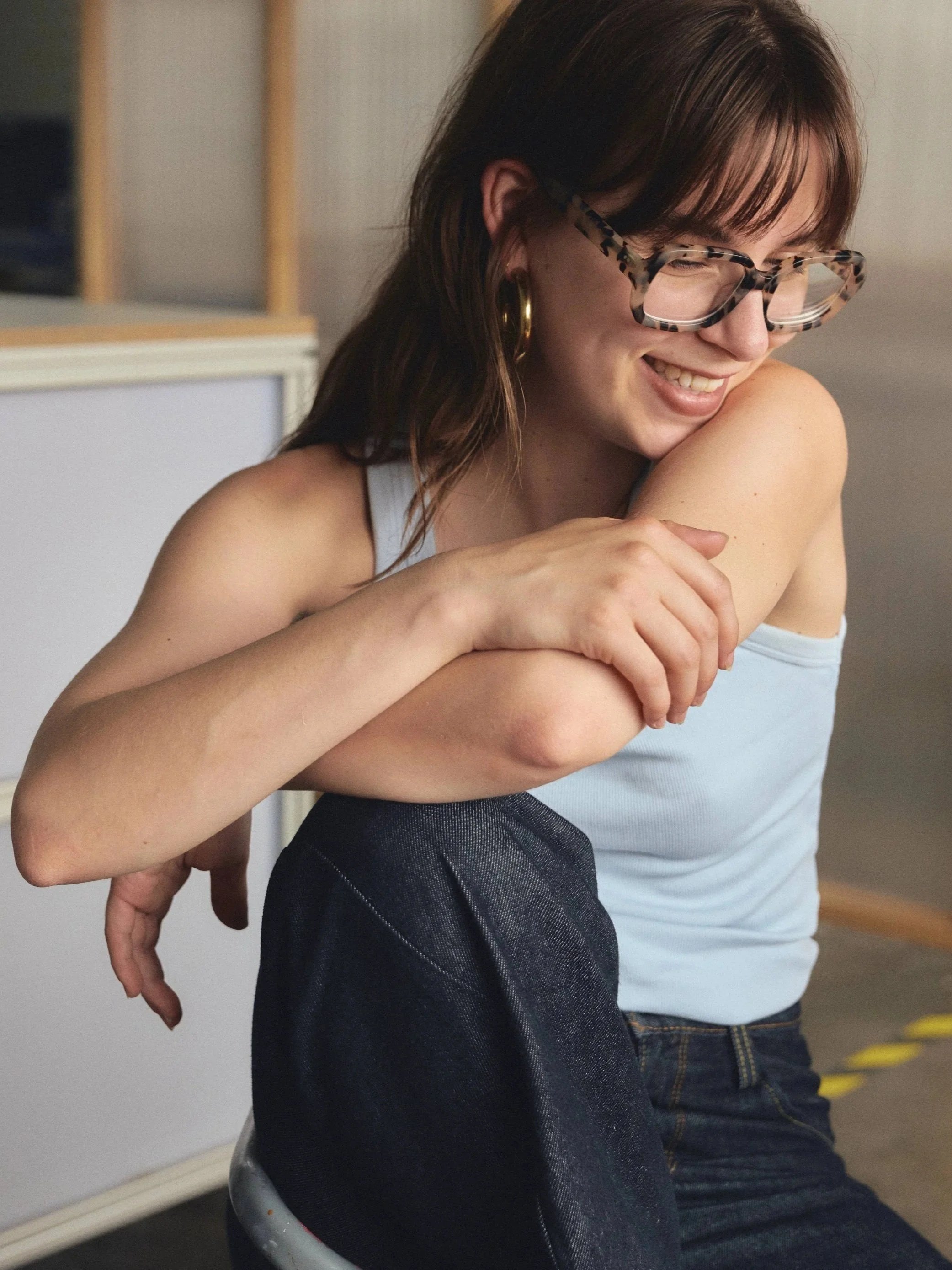 A woman with brown hair, wearing glasses with a leopard print pattern and yellow earrings, laughing and leaning over a child, who is sitting in a gray chair, with her arm draped over the child's shoulder.