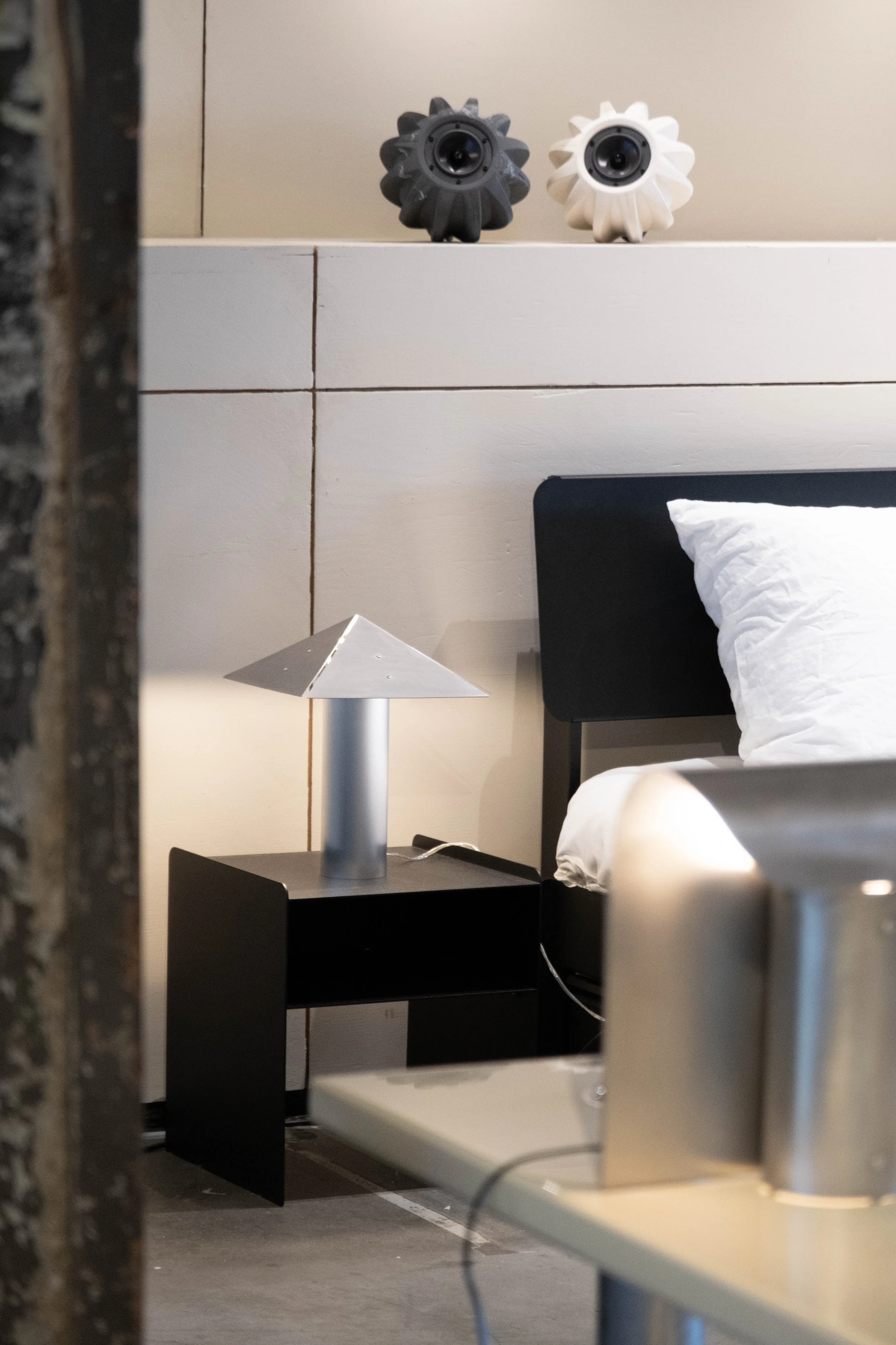 View of a black bedside table with a modern table lamp, white bedsheet and pillow, and two decorative objects resembling sea urchins on the wall behind.