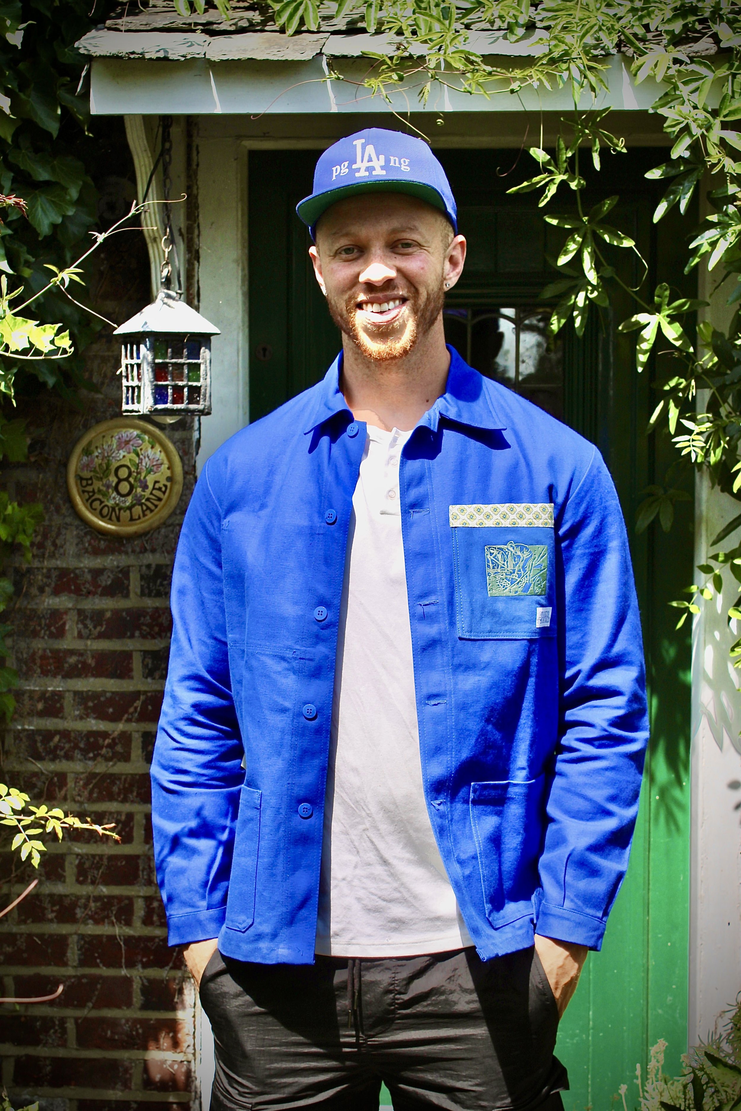 A man standing outdoors, smiling, wearing a blue LA Dodgers cap, a bright blue shirt with patches, and dark shorts. There are green plants and a decorative lantern hanging nearby, with a sign on the wall reading '8 Bacon Lane' in the background.