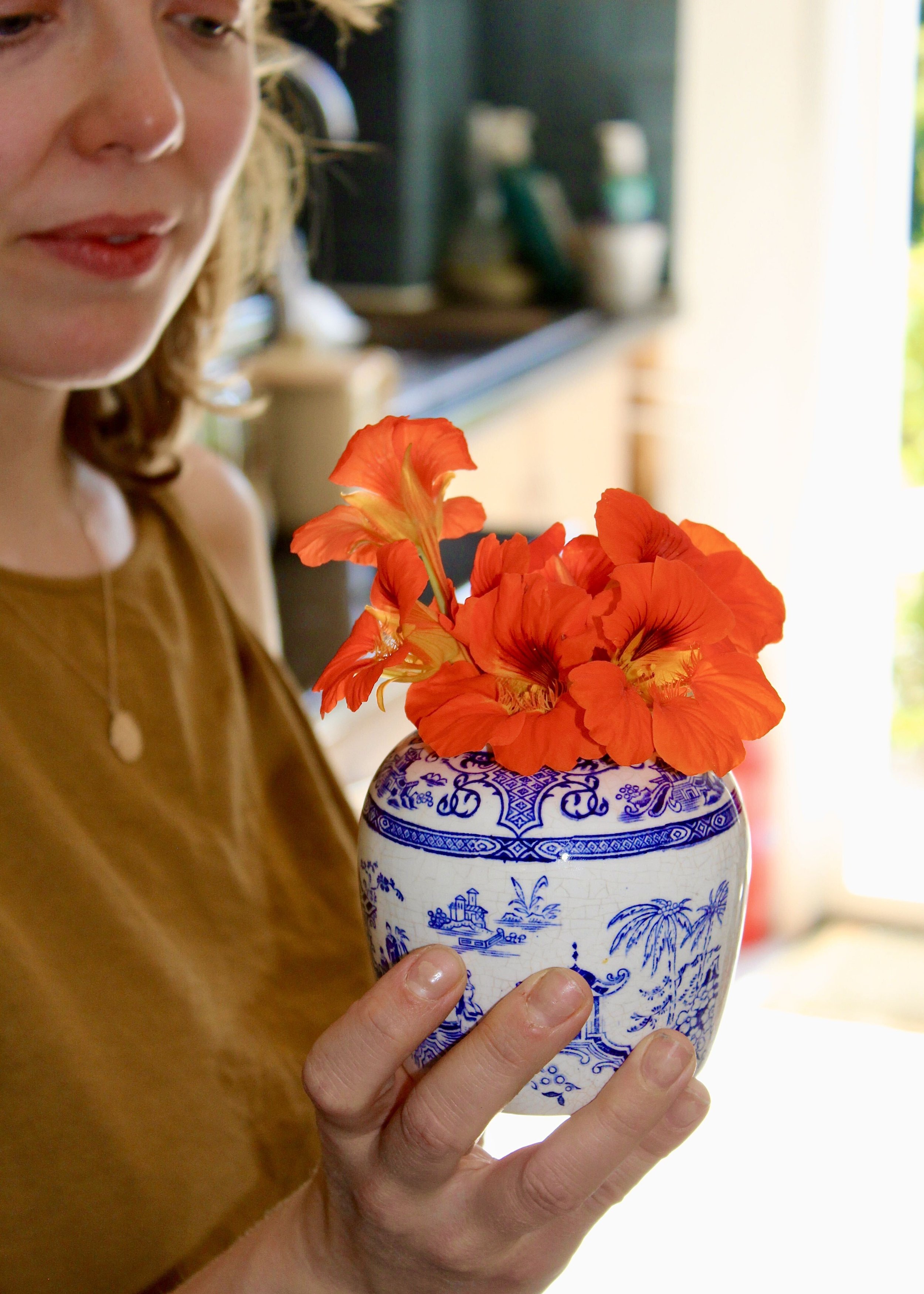A person holding a blue and white porcelain vase with orange flowers inside, with only part of their face and hand visible.