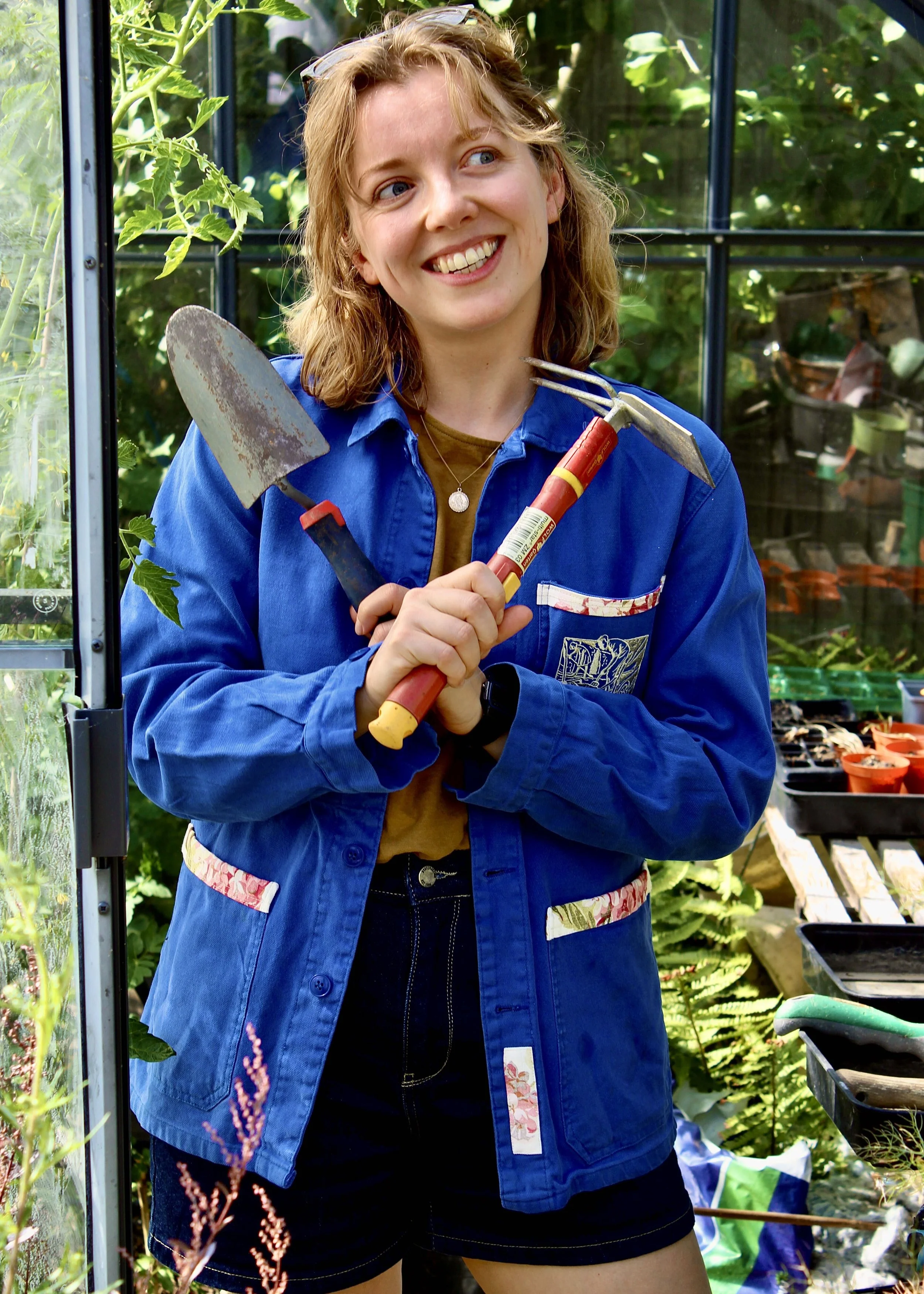 A young smiling woman with blonde hair and blue eyes, wearing a blue jacket and holding gardening tools, standing inside a greenhouse surrounded by lush green plants.
