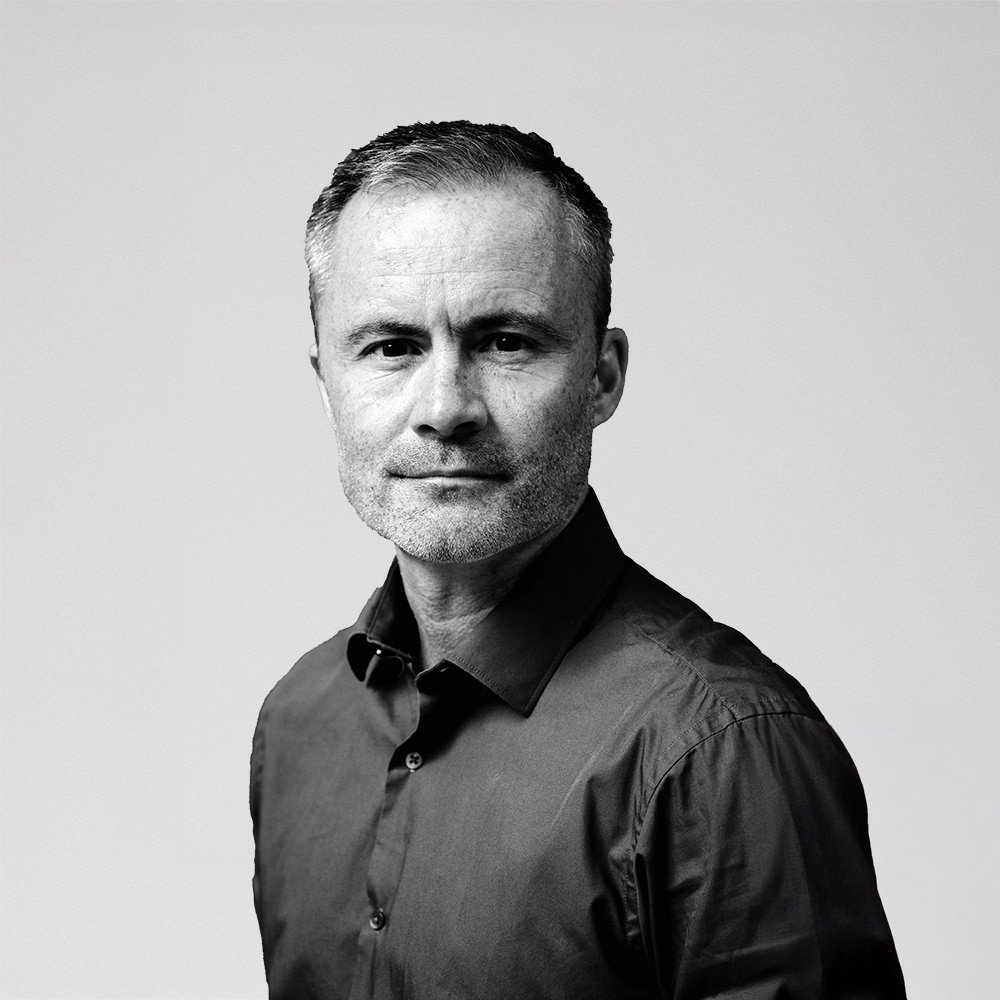 Black and white portrait of Ben Verinder wearing a collared shirt, looking directly at the camera against a plain background.