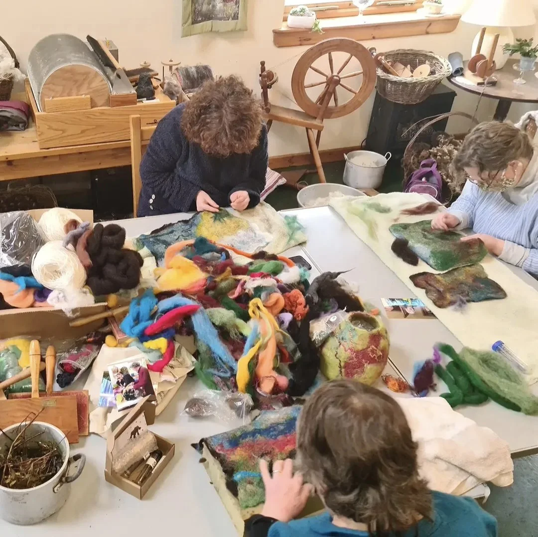 People working on colorful felted wool projects at a craft workshop table, surrounded by wool and felting supplies.