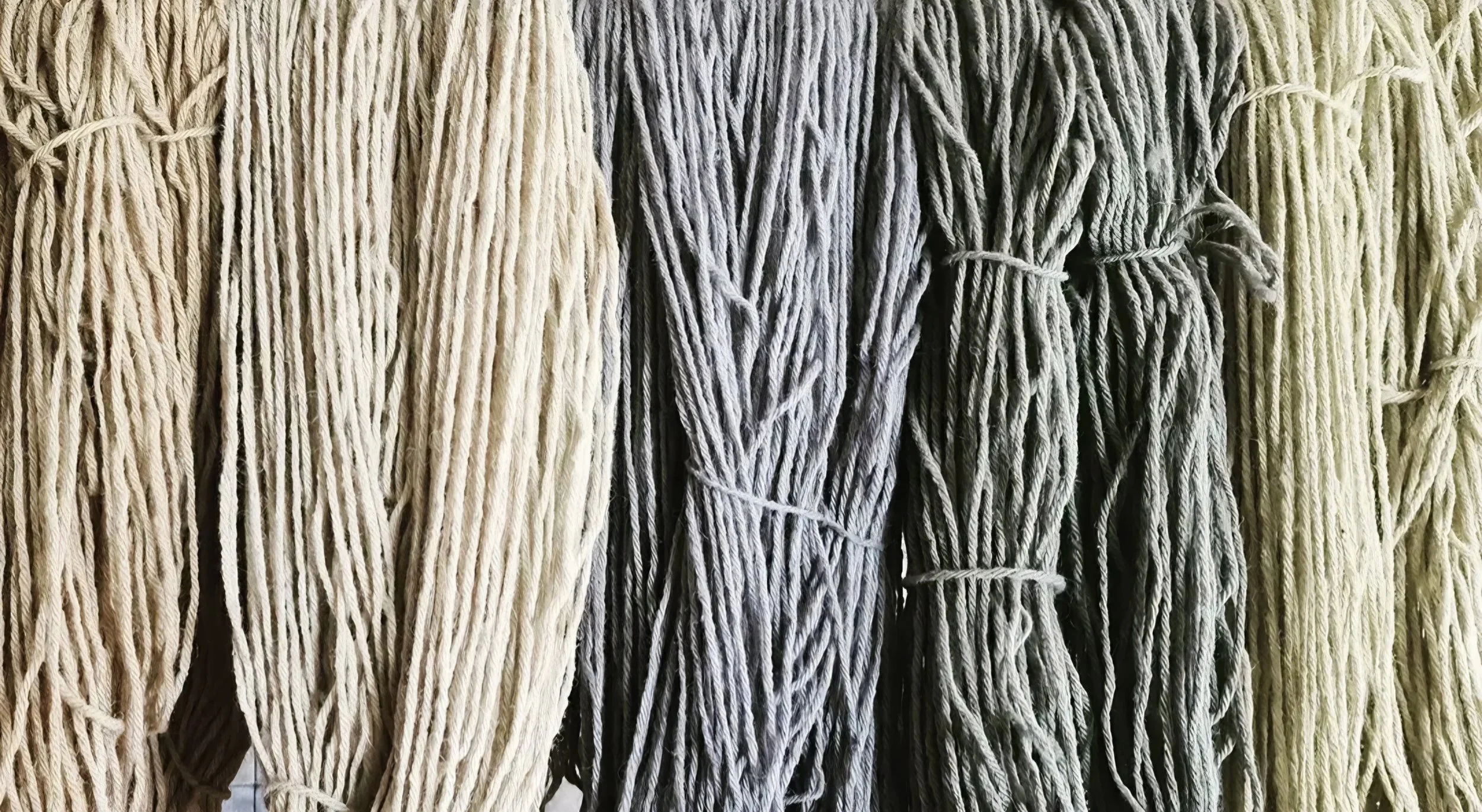 Bundles of naturally dyed yarn in white, gray, and beige yarn or rope hanging side by side.