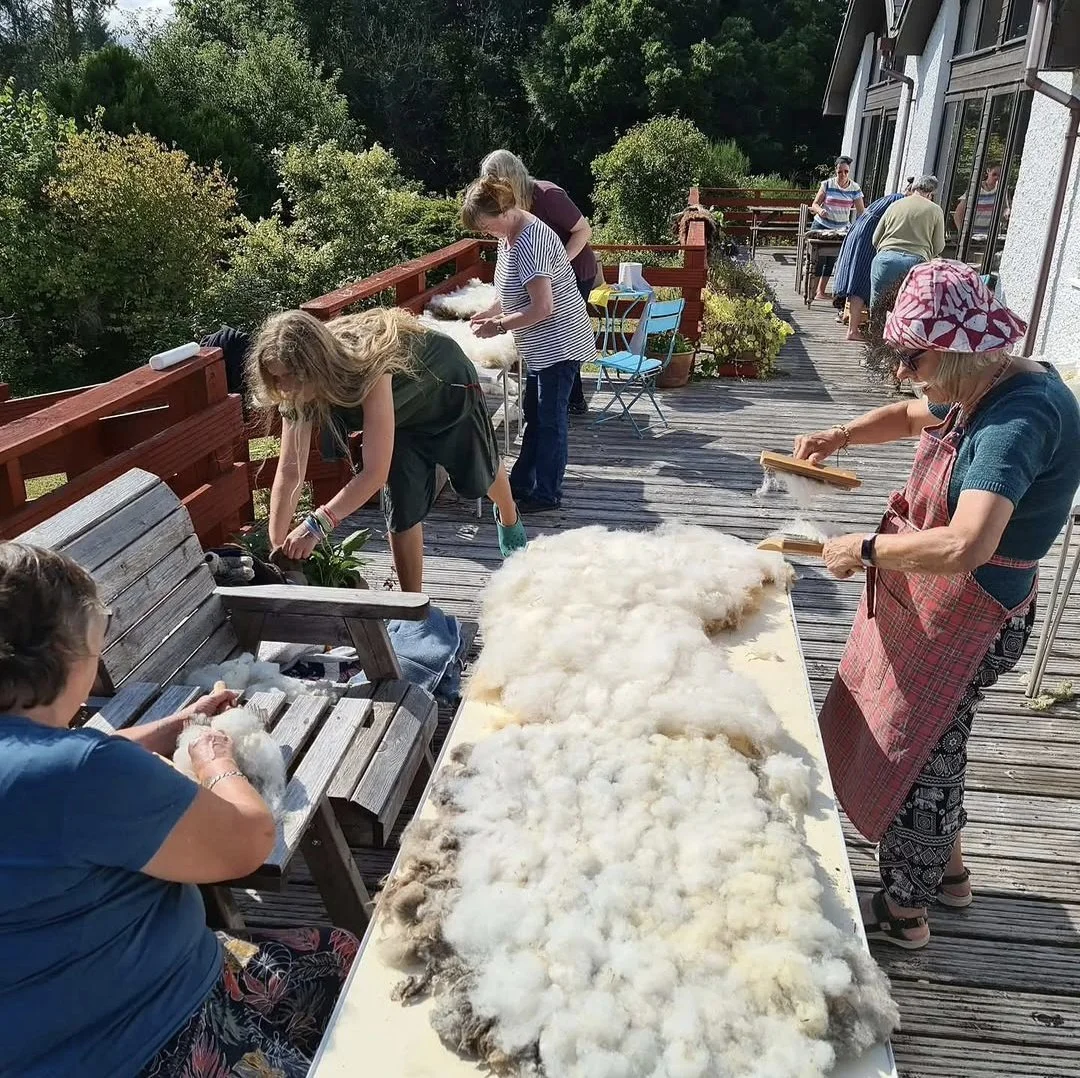 People gathering outdoors on a wooden deck, working with wool fibers, possibly shearing or processing wool, with tables piled with raw wool and various tools, surrounded by greenery.