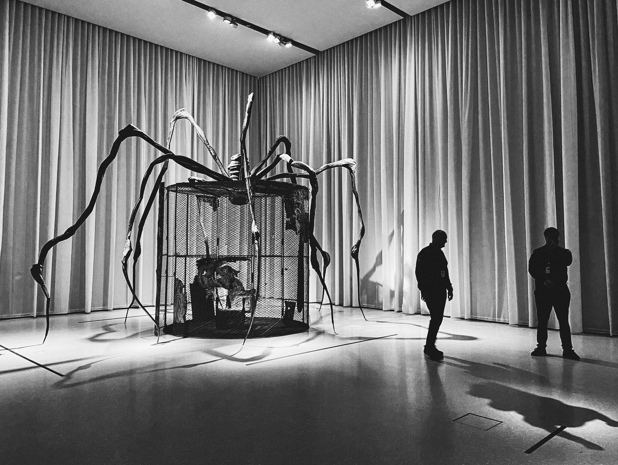 A large, spider-like sculpture with long, twisted legs extends over a small enclosed space, with two security guards standing nearby in a room with tall curtains.