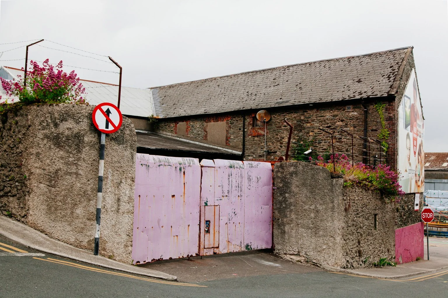 An old brick building with a slanted roof, pink metal gate, and a pink lower wall. There is a no-entry traffic sign near it, and a stop sign at the street corner.