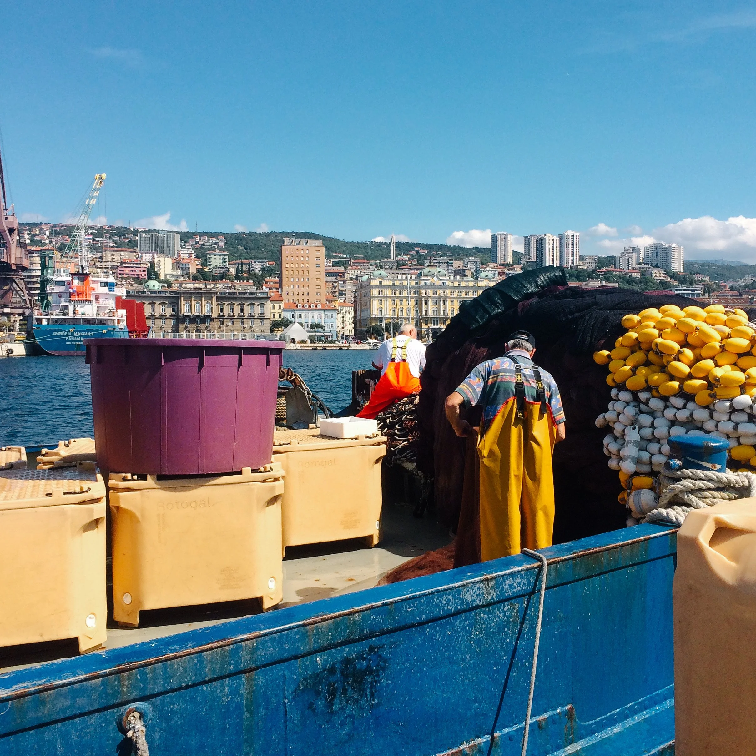 Marine workers on a dock surrounded by fishing gear, with a city skyline and water in the background.