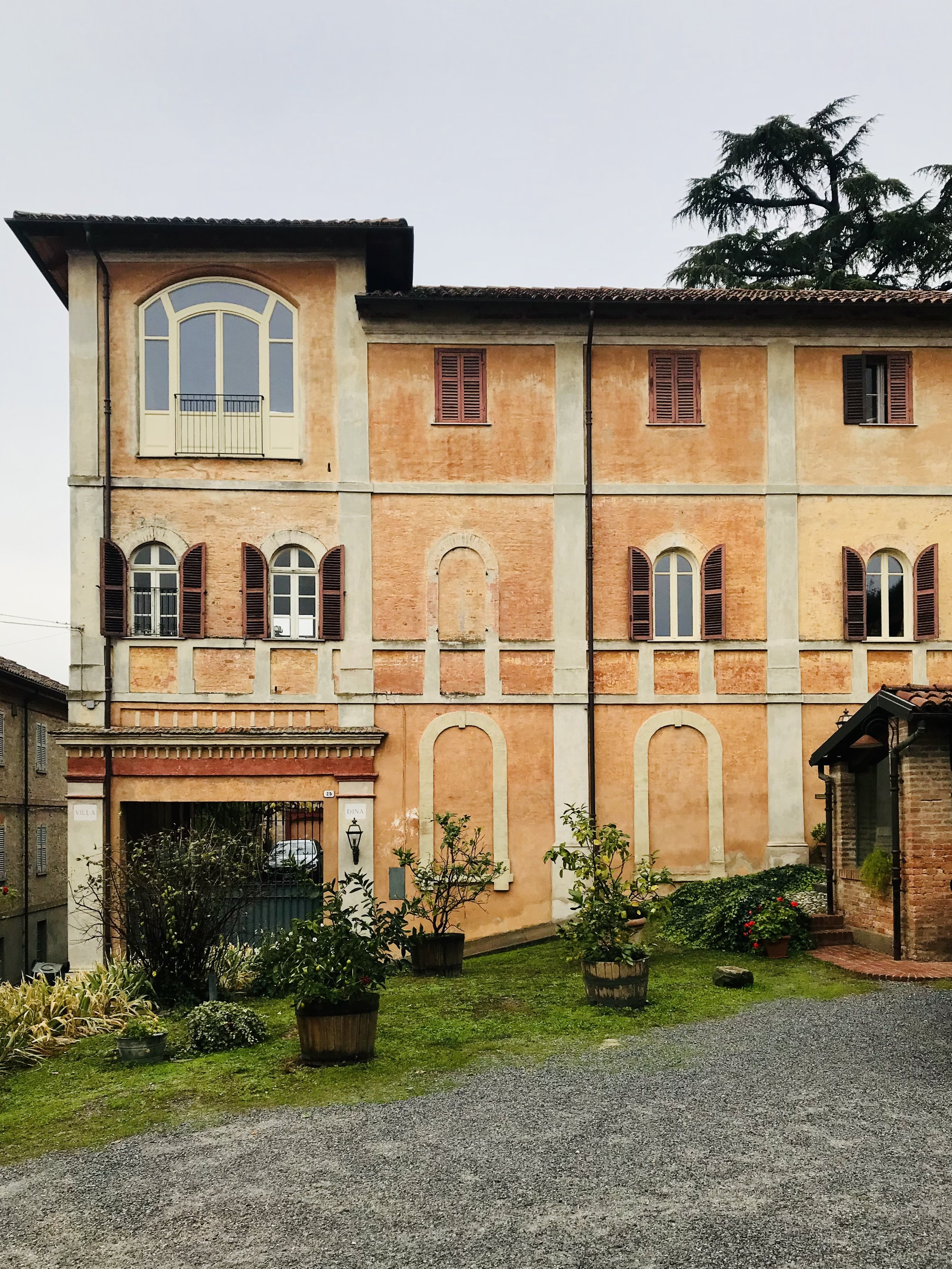 Edificio storico in mattoni con finestre ad arco e persiane marroni, ingresso con cancello e giardino con piante e cespugli.