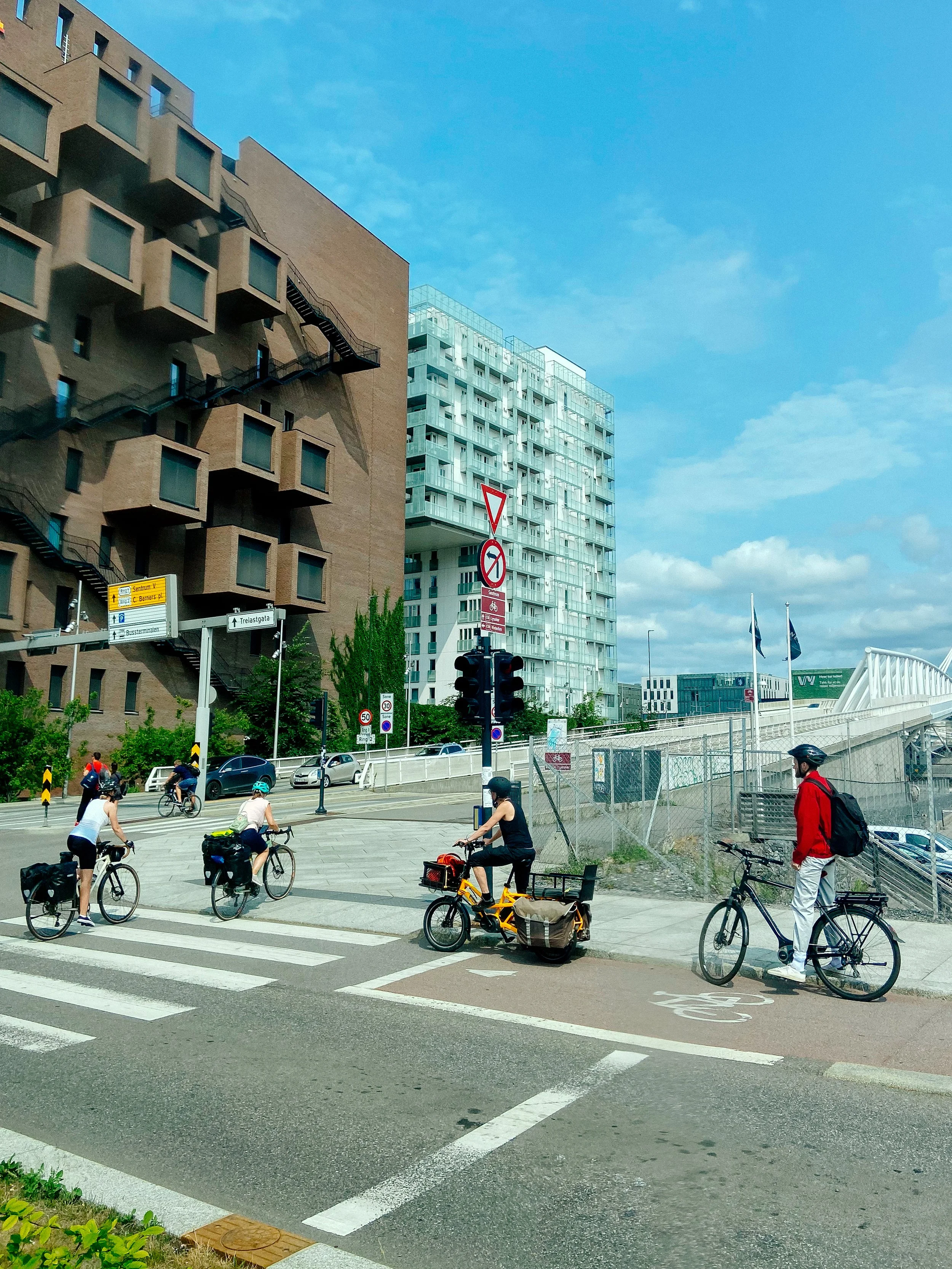 Group of cyclists waiting at a crosswalk in an urban area with modern buildings, clear blue sky, and traffic signals.