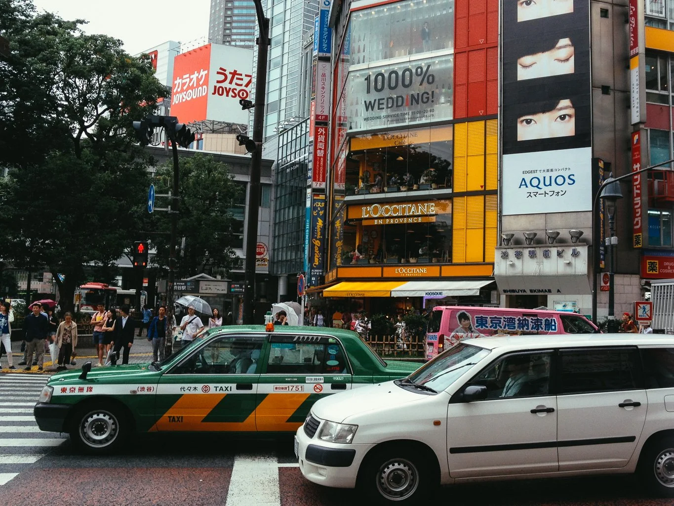 City street scene with taxis and pedestrians, tall buildings with colorful advertisements including L'Occitane store, and a large billboard with Japanese characters.