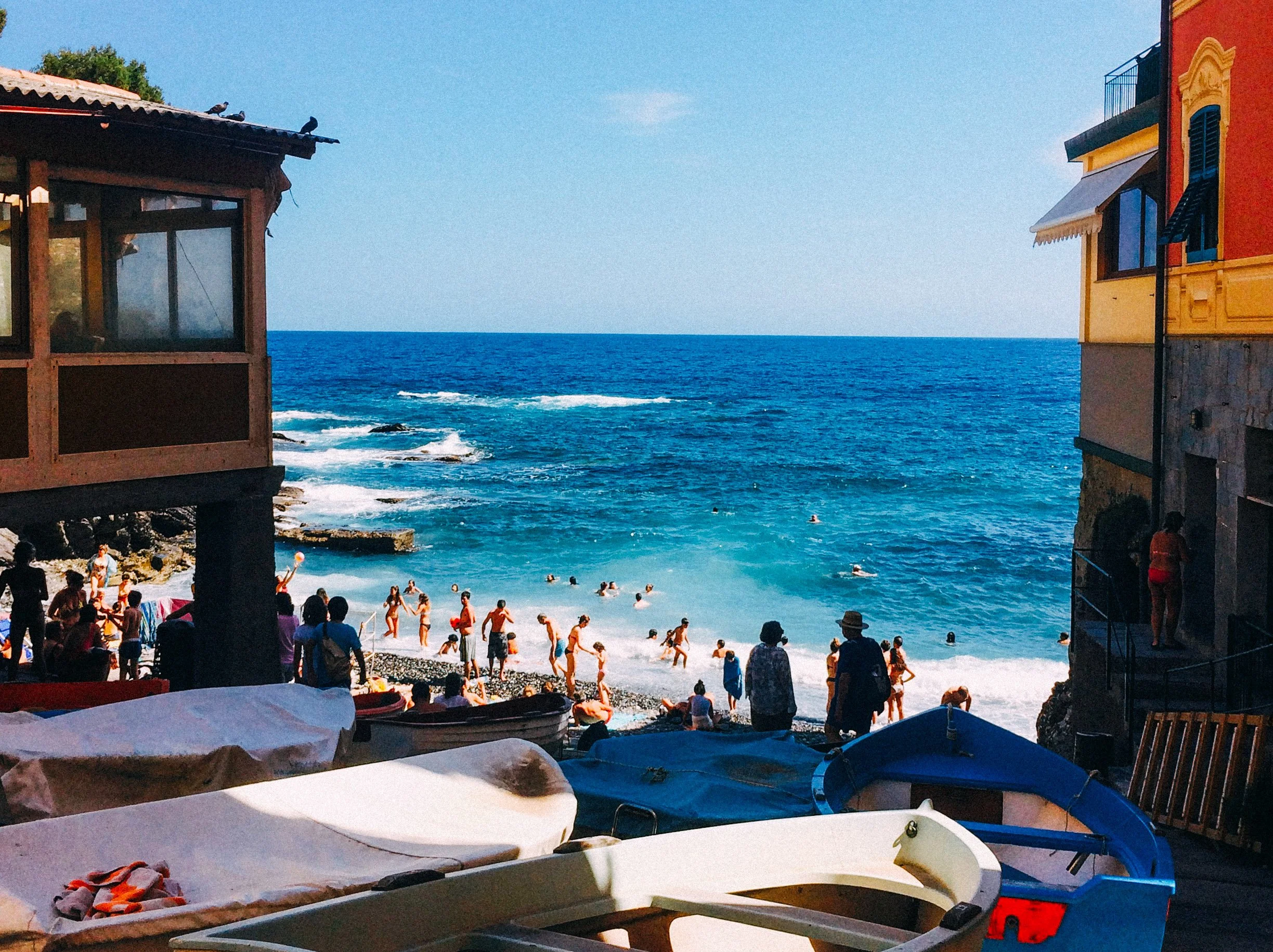View from a beachside cafe showing a lively beach with people swimming and playing in the water, colorful buildings on either side, lounge chairs and boats in the foreground, ocean waves, and a clear blue sky.