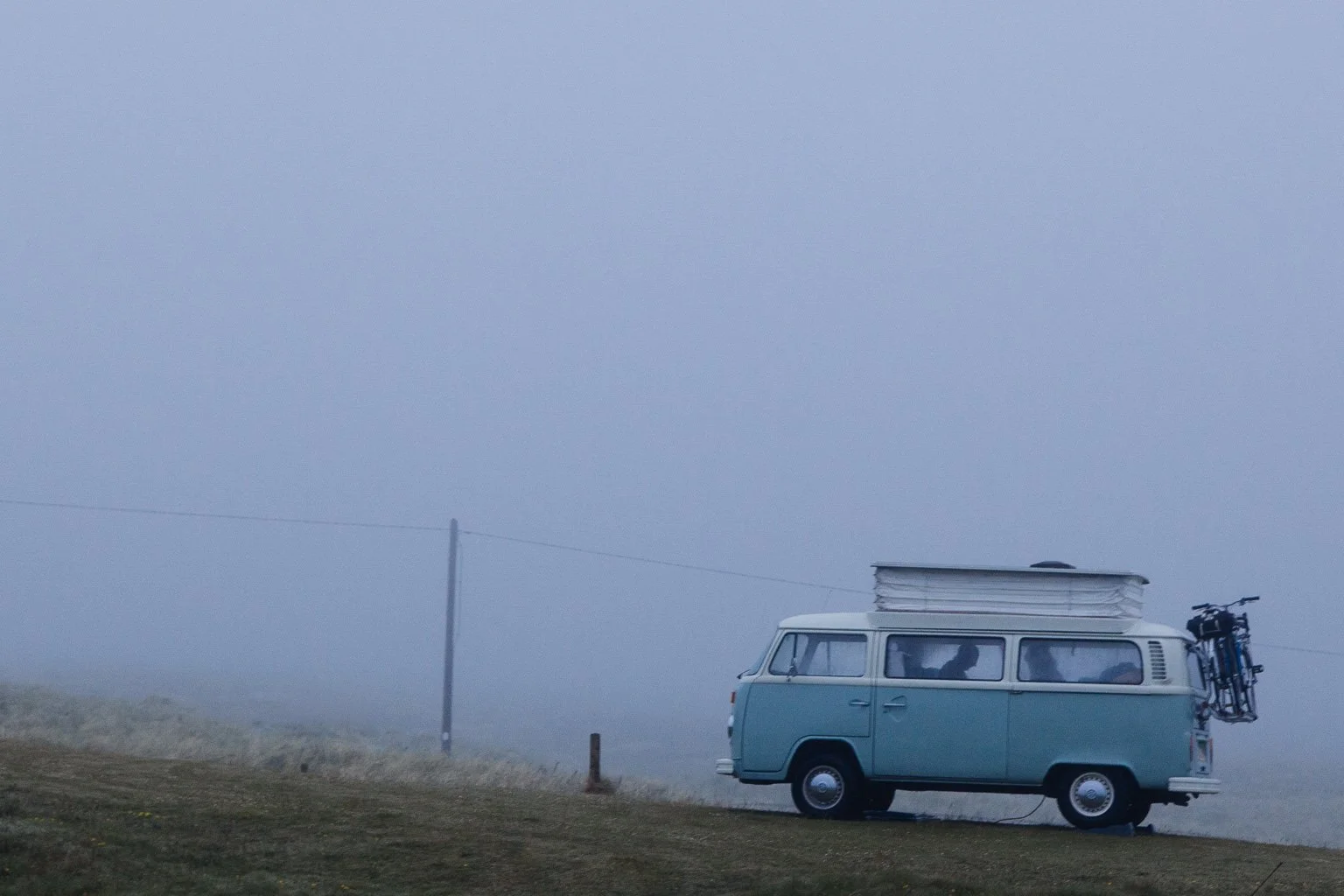 A vintage blue van parked on a grassy field with a pop-up roof and a bicycle mounted on the rear, shrouded in foggy weather.