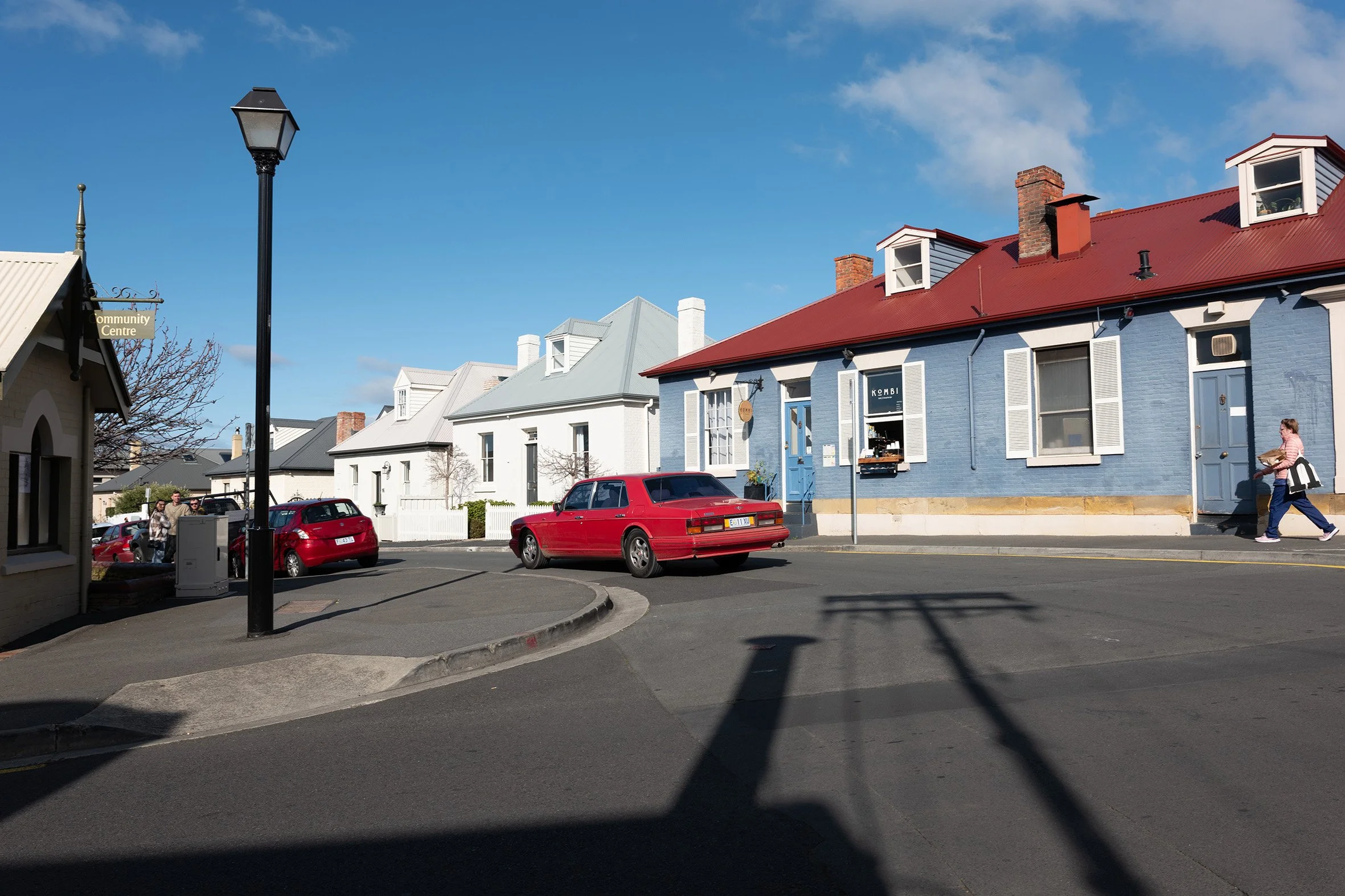 A street scene with a red car parked on the corner, white and blue houses in the background, a streetlamp, and a woman carrying a bag walking past a blue house.