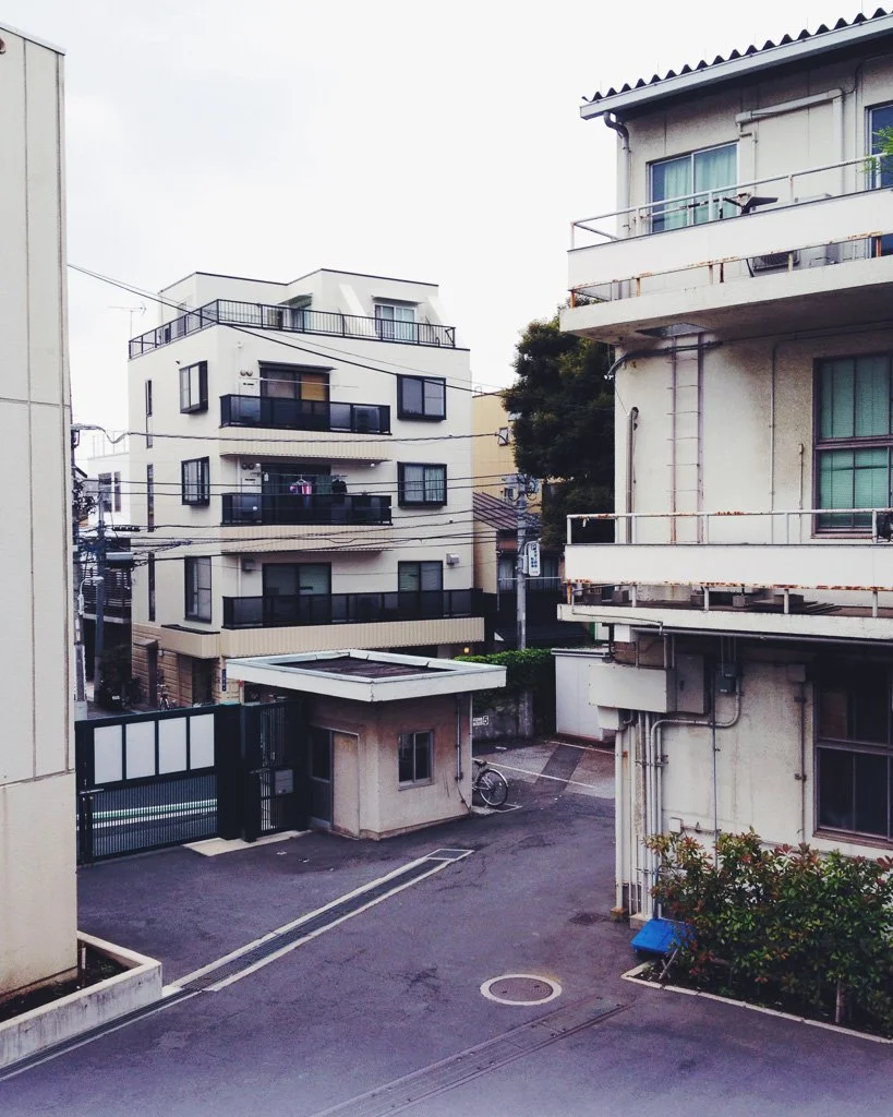 View of a residential area with multiple apartment buildings, a small guardhouse at a gated entrance, a bicycle parked near the guardhouse, and overhead power lines on an overcast day.