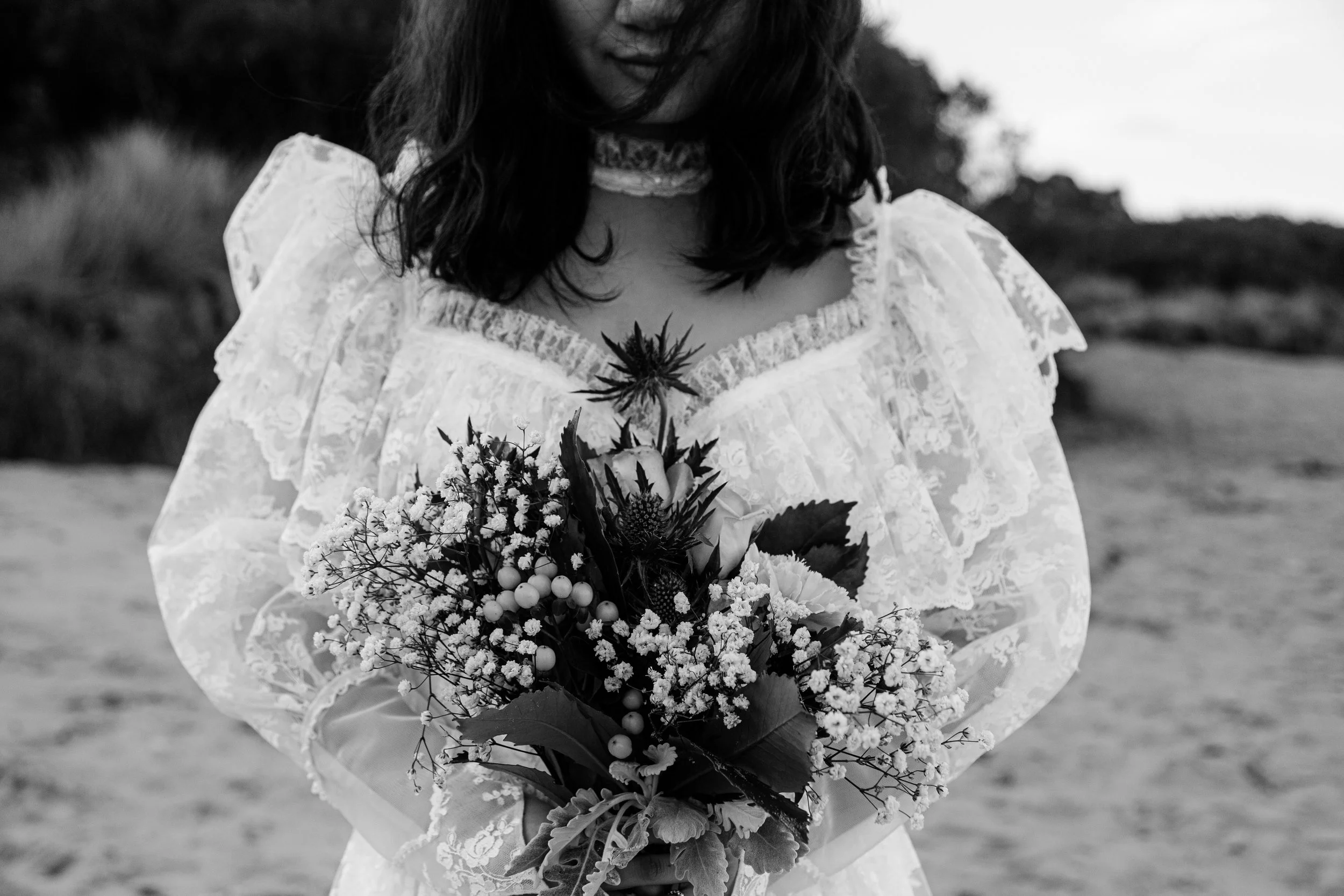 Black and white photo of a woman in a lace dress holding a bouquet of flowers outdoors, with trees and sky in the background.