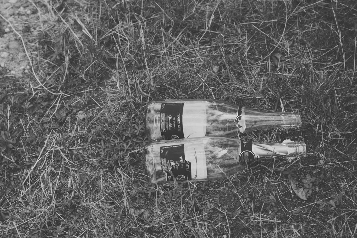 Two broken bottles of Trento brandy lie on the ground among grass and leaves.