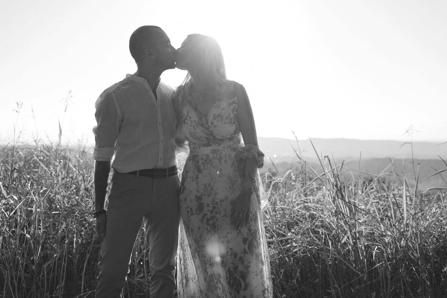 A couple kissing in a field with tall grass, backlit by the sun, in black and white.