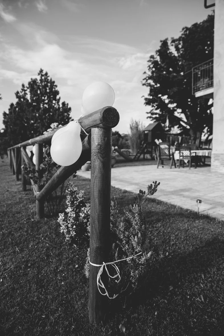 A wooden fence decorated with balloons and greenery outside a house with patio furniture, and trees in the background.