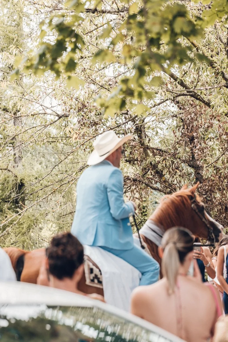 Person in a light blue suit and wide-brimmed hat riding a horse amidst trees, with people around.