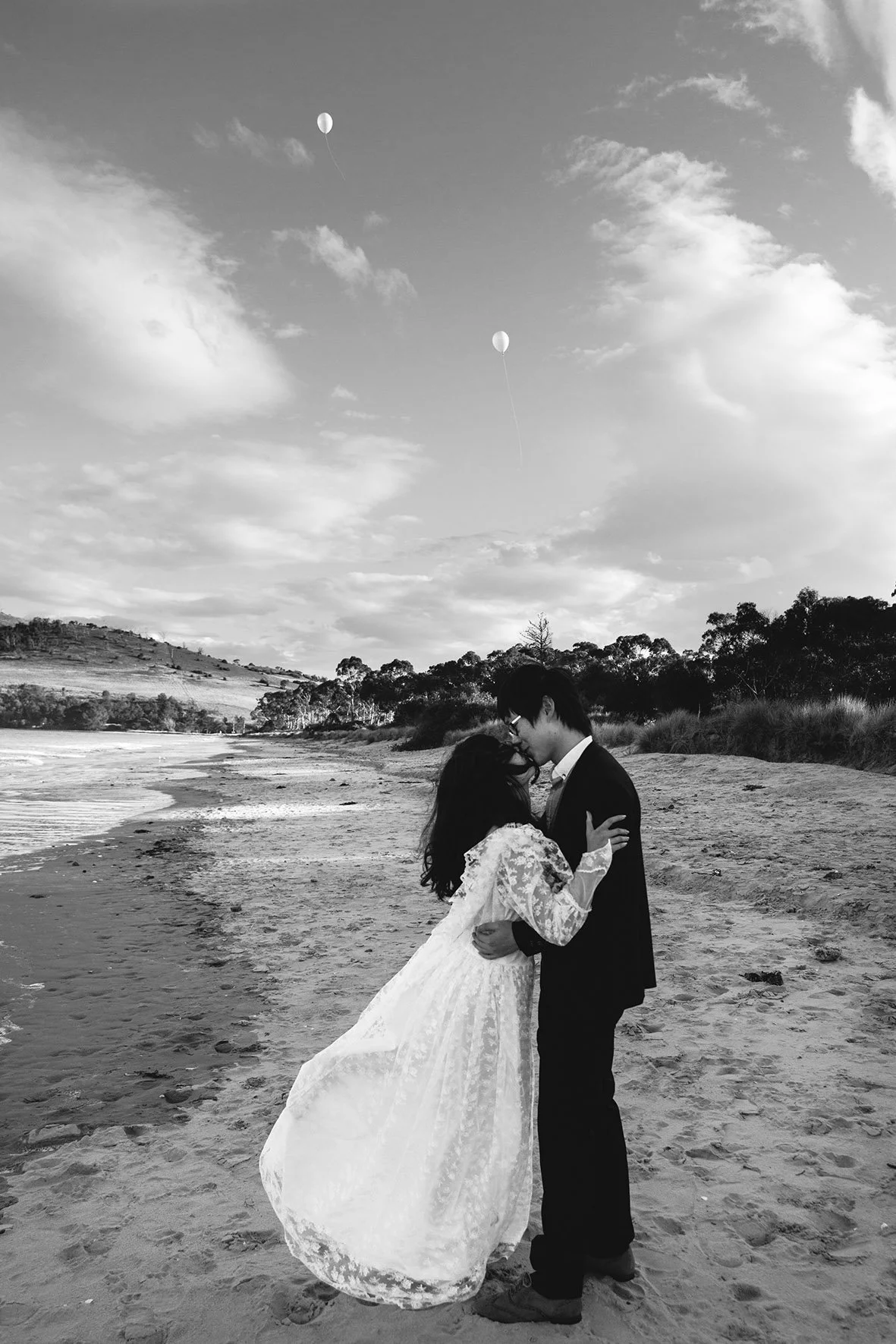 A couple in wedding attire embracing on a beach with two helium balloons in the sky.