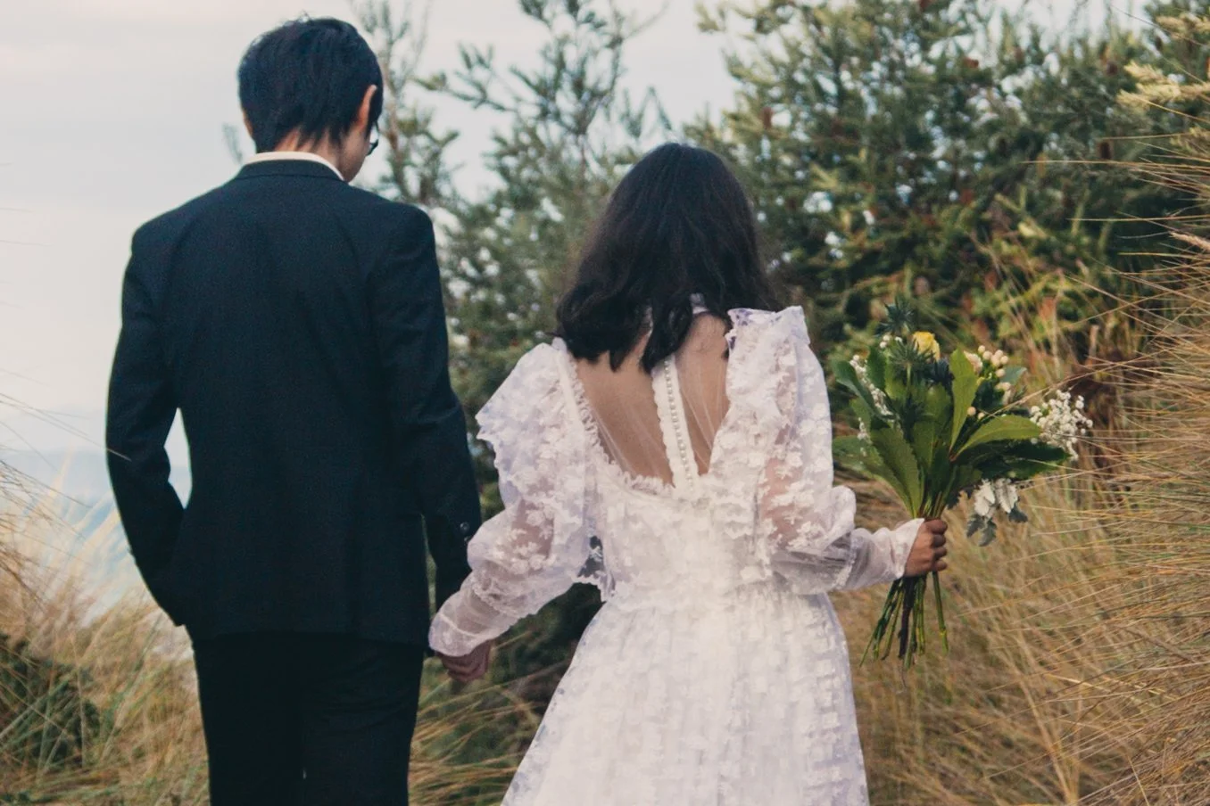 A bride and groom hold hands and walk through a grassy outdoor area, the bride carrying a bouquet of flowers, during a wedding photoshoot.