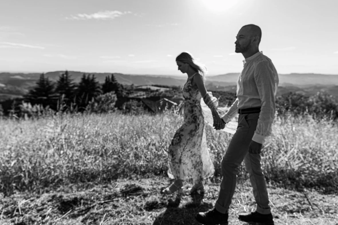 A black and white photo of a man and woman holding hands while walking through a grassy field during sunset, with mountains and trees in the background.